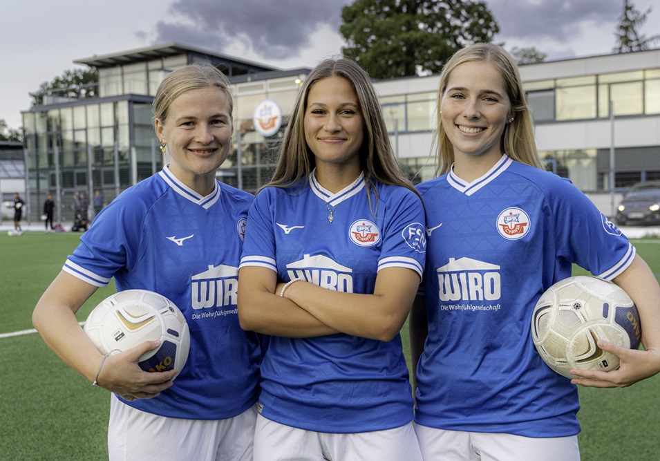 Drei junge Fußballerinnen im blauen Hansa-Trikot mit dem Logo der WIRO darauf.