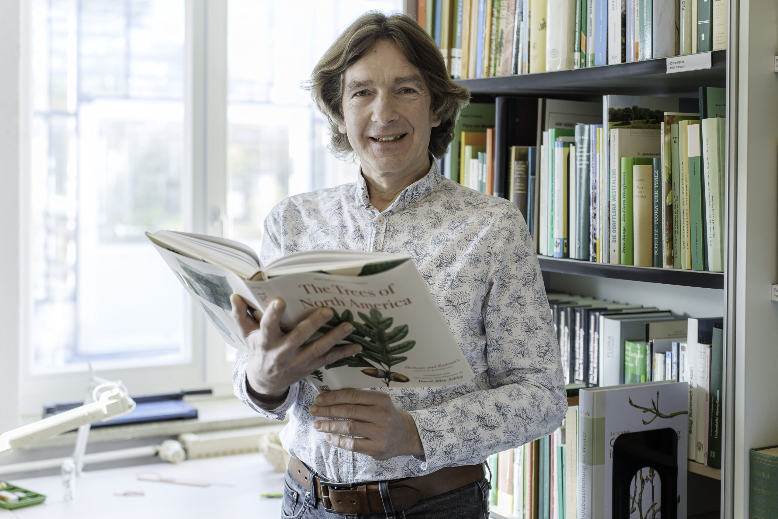 Dr. Dethardt Götze, Kustos des Botanischen Gartens, braune längere Haare, steht vor einem Regal mit Fachliteratur