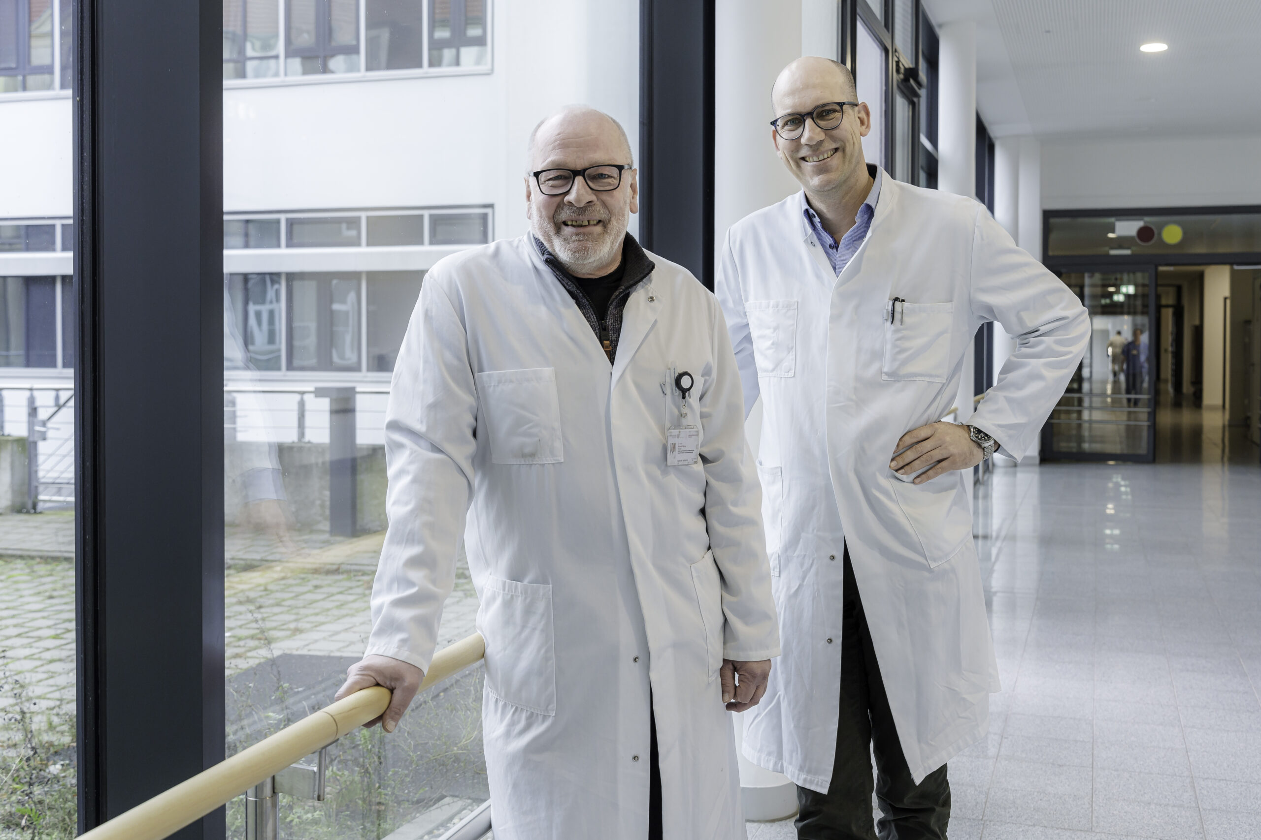 Doktor Ronald Siems (li.) und Professor Sebastian Hinz, beide kahlköpfig und mit schwarzen Brillen, stehen im weißen Kittel im verglasten Flur des Krankenhauses. FOTO: DOMUSIMAGES