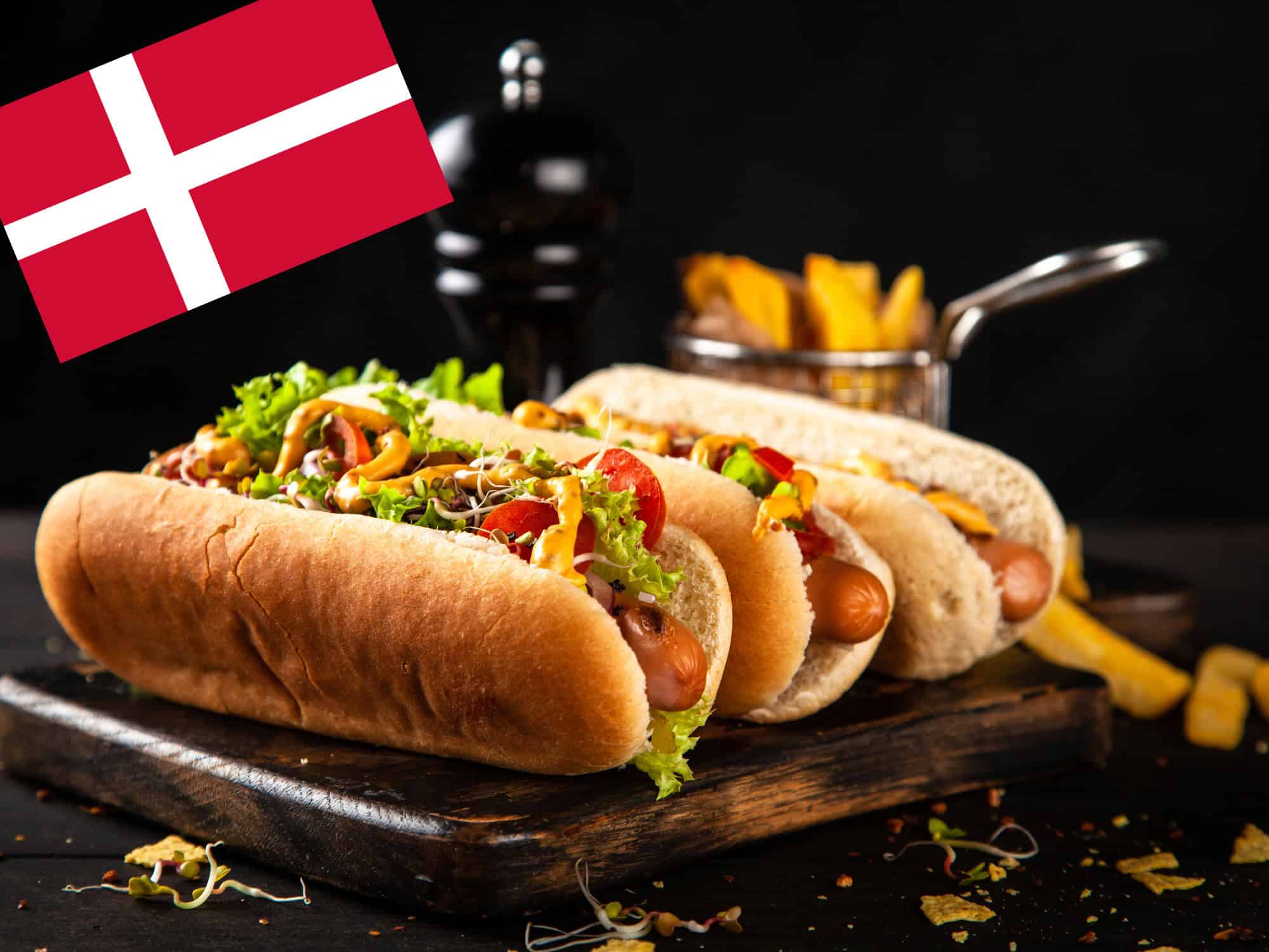 25-02-25 Hotdog Bild Serviervorschlag drei Hot Dogs auf einem dunklen Holzbrett, im Hintergrund ein Pfefferstreuer und Pomm Frites, oben links eine dänische Flagge