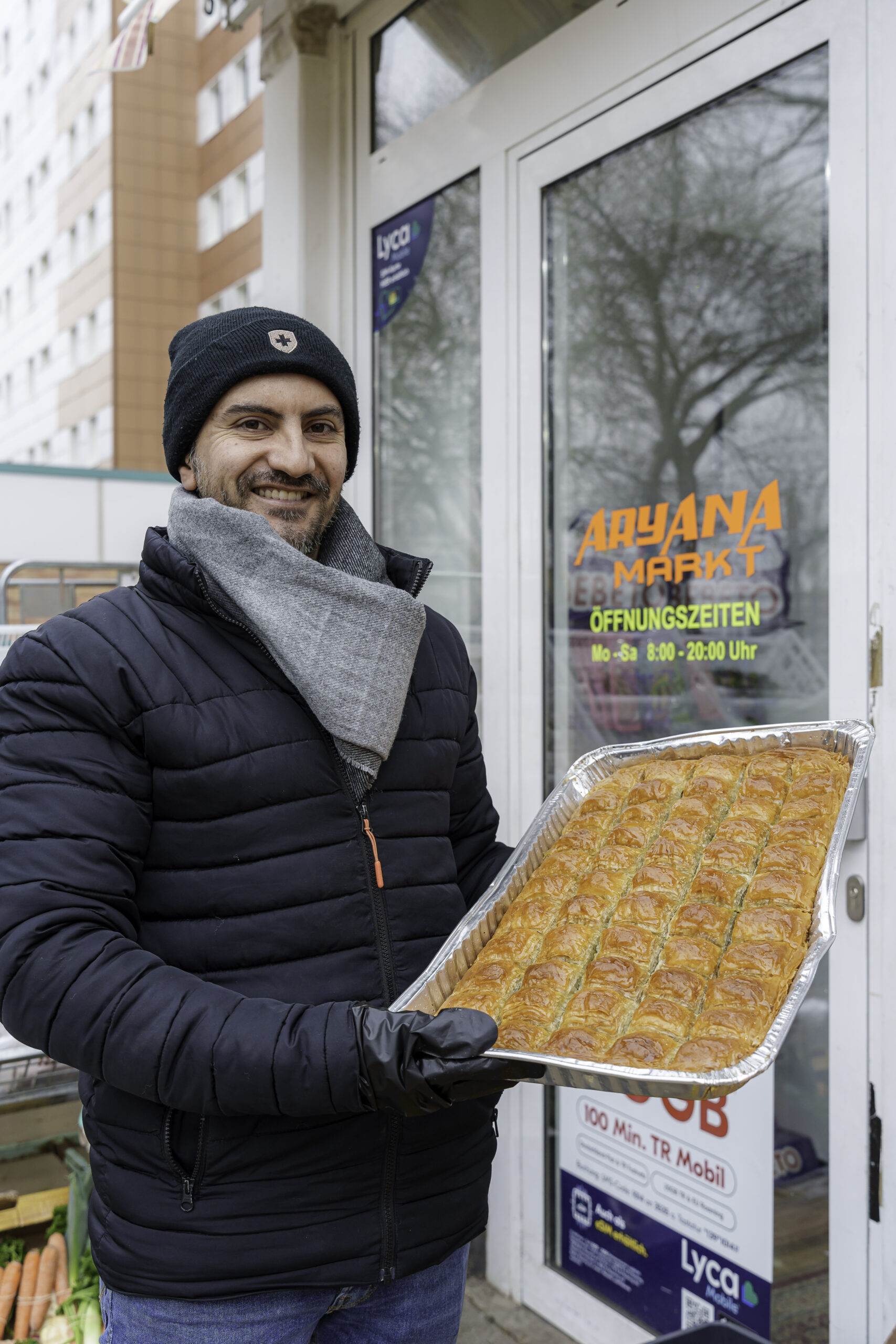 Der Inhaber des Aryana Marktes mit dunkler Mütze und Jacke mit einem gefüllten Backblech vor seinem Geschäft.