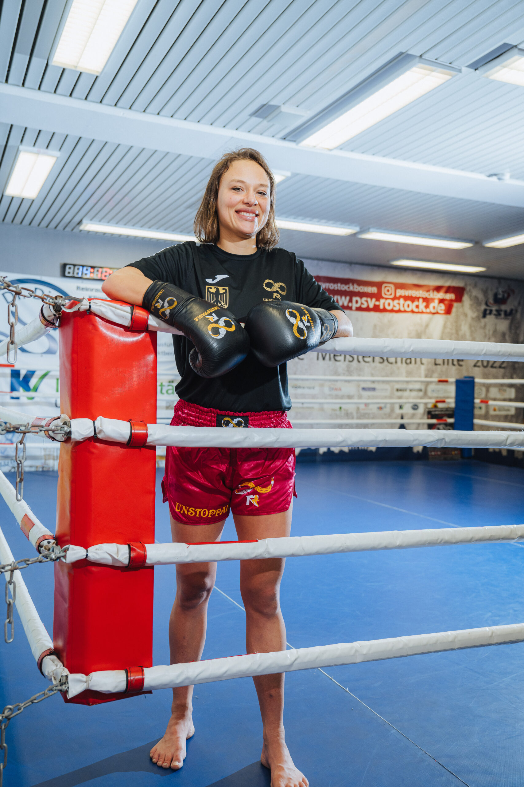 Jette Körner, braune lange Haare, lachend, steht im Ring. Sie trägt ein schwarzes Shirt und rote kurze Sport-Shorts.