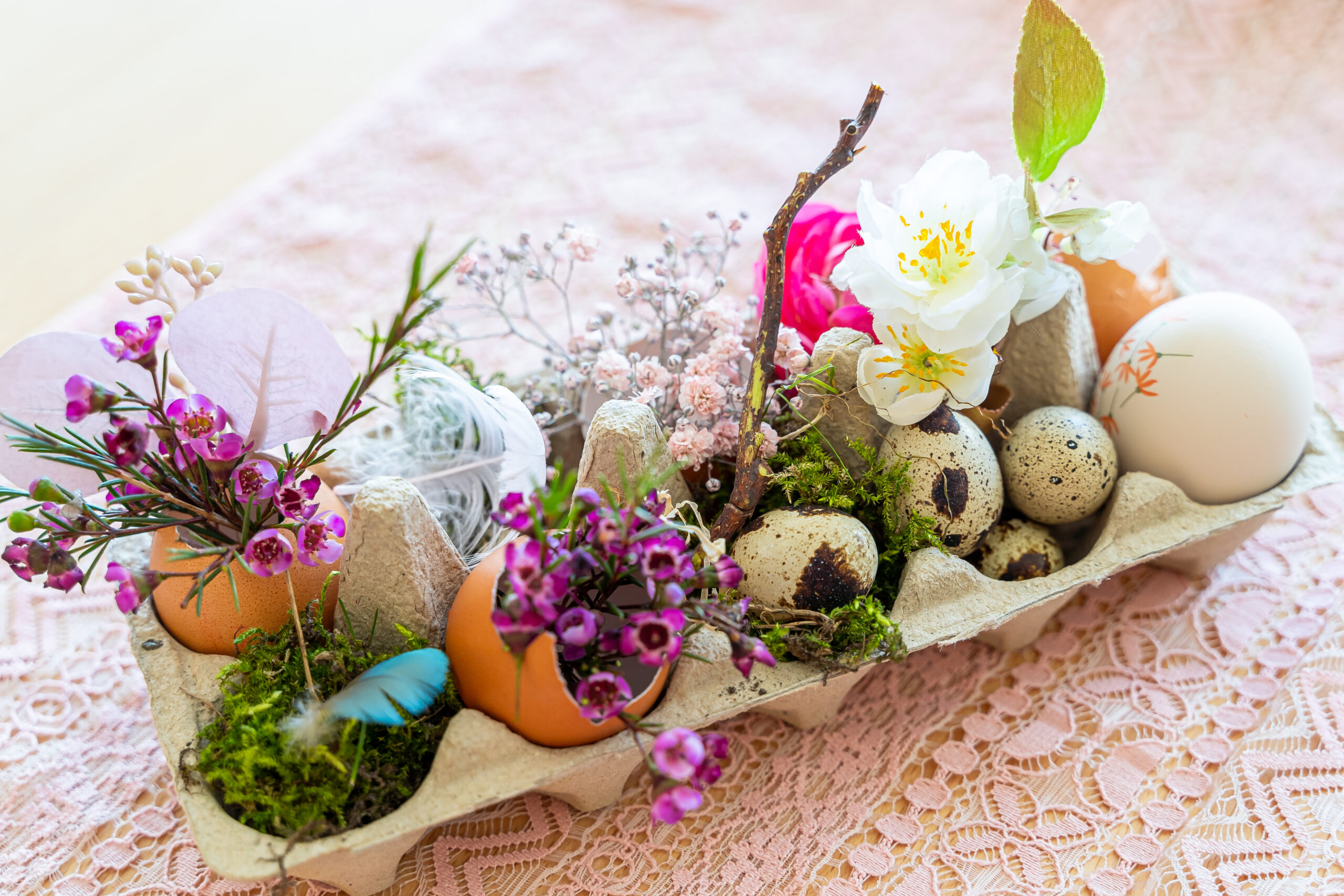 Ein Osternest mit Moos, Federn, Eiern, weißen, pinken und rosa Blüten, verziert mit grünem Blatt und einem Stöckchen.