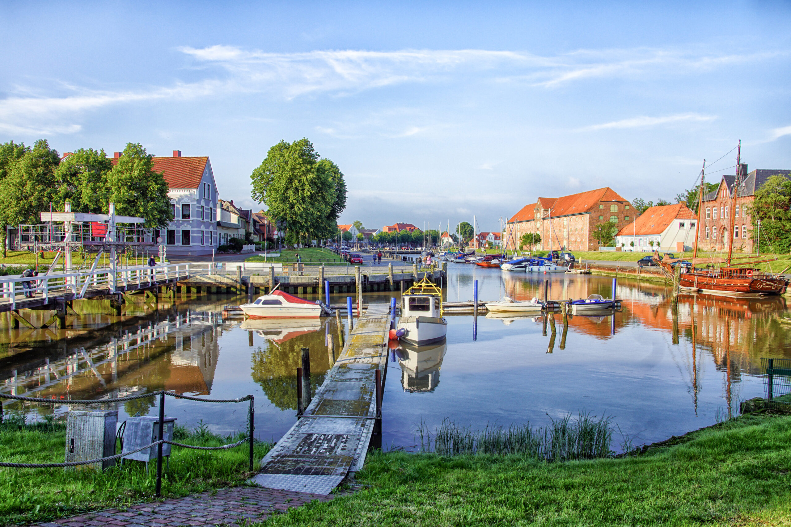 Der Hafen von Tönning: grasbewachsenes Ufer, rote Backsteinhäuschen, kleine Bote auf blauem Wasser.