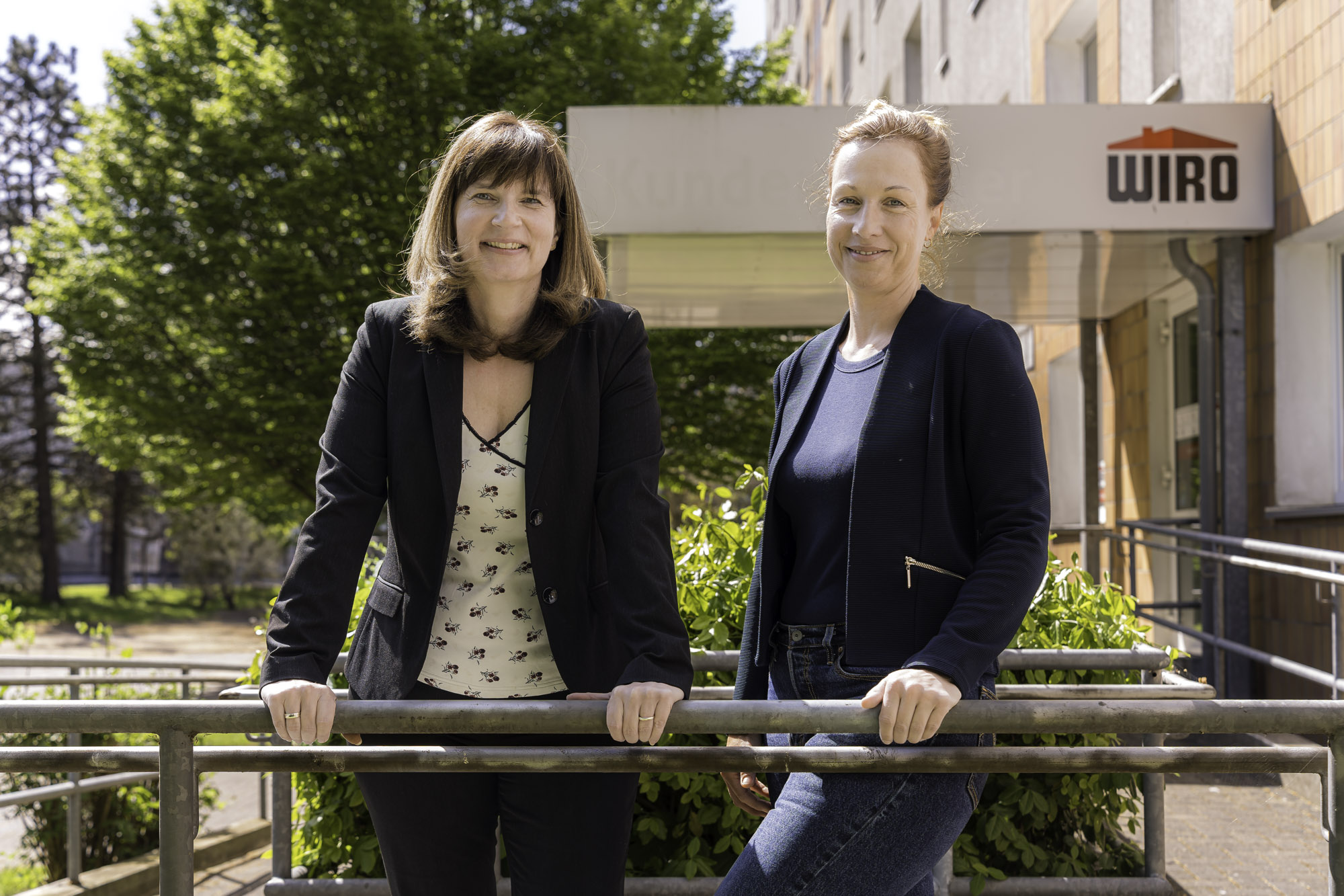 Silvana Stephanski und Katharina Krüger lehnen sich entspannt an das Treppengeländer des WIRO-KundenCenters Forderungs- und Sozialmanagement. Im Hintergrund das Eingangsschild "WIRO" und ein voller grüner Baum.