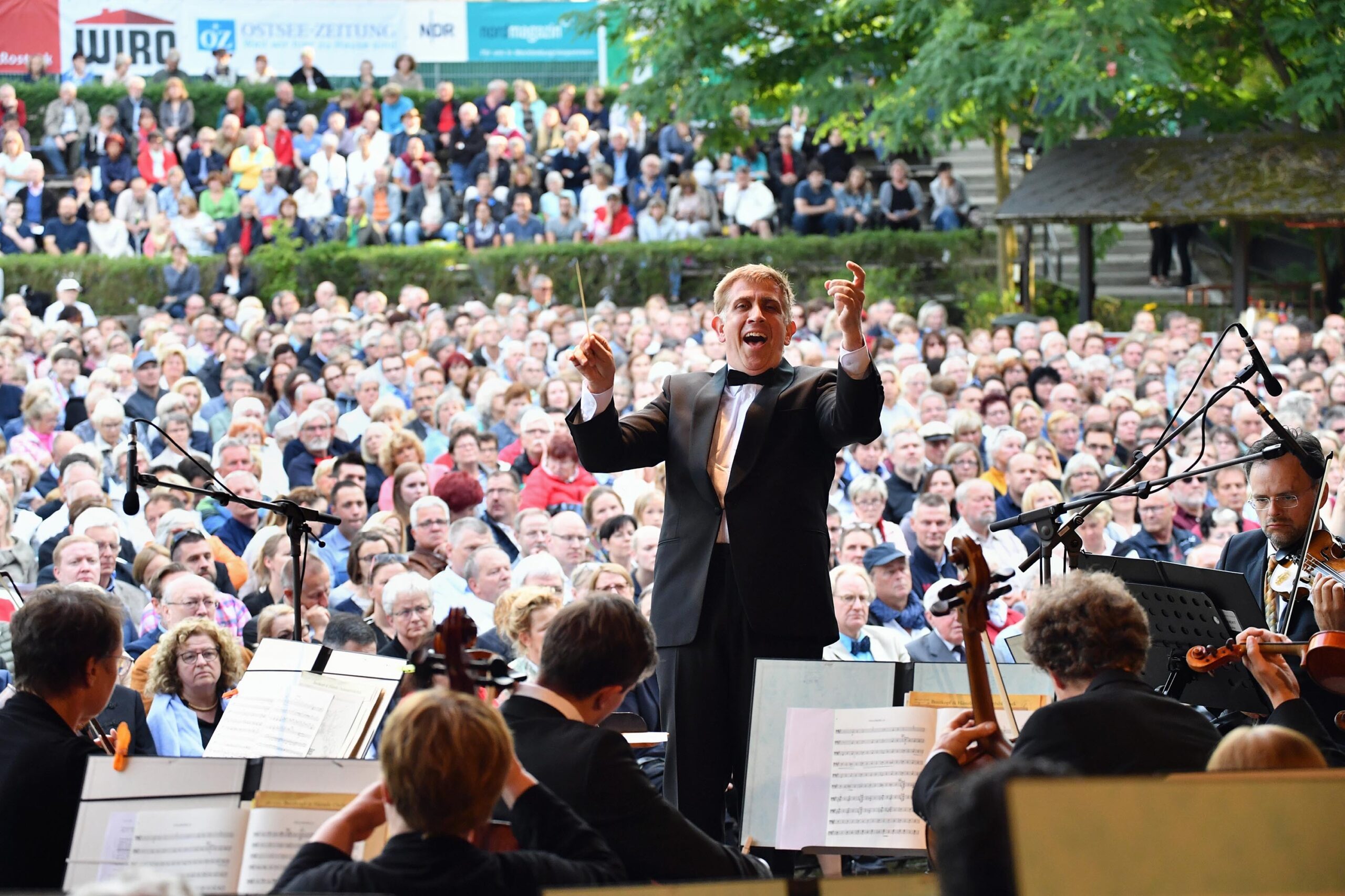 Ein Dirigent hebt den Taktstock, Musiker spielen Instrumente, das Publikum lauscht.