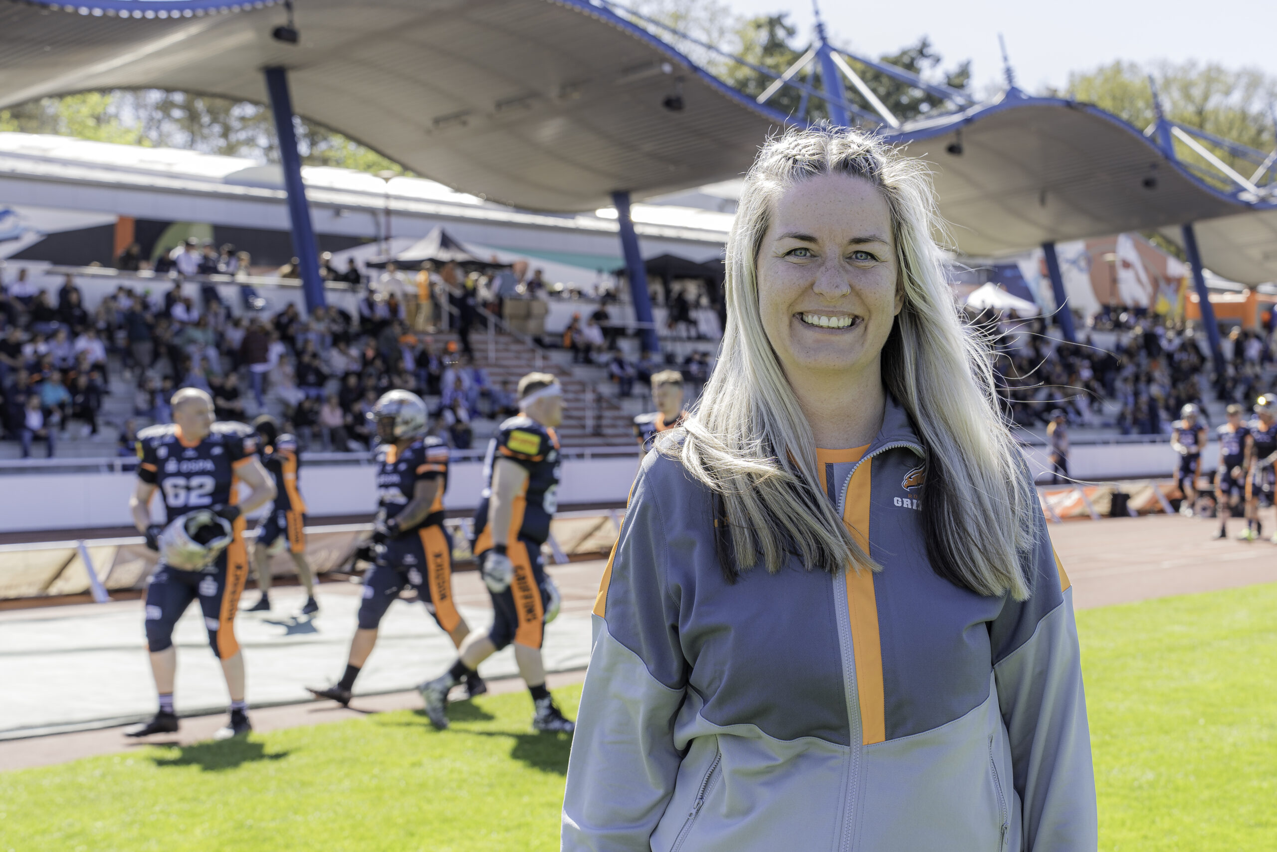 Peggy Schröder von den Rostocker Griffins in grau-orangefarbenem Trainingszeug lacht in die Kamera, im Hintergrund sind die Spieler der Griffins beim Aufwärmen zu erkennen.