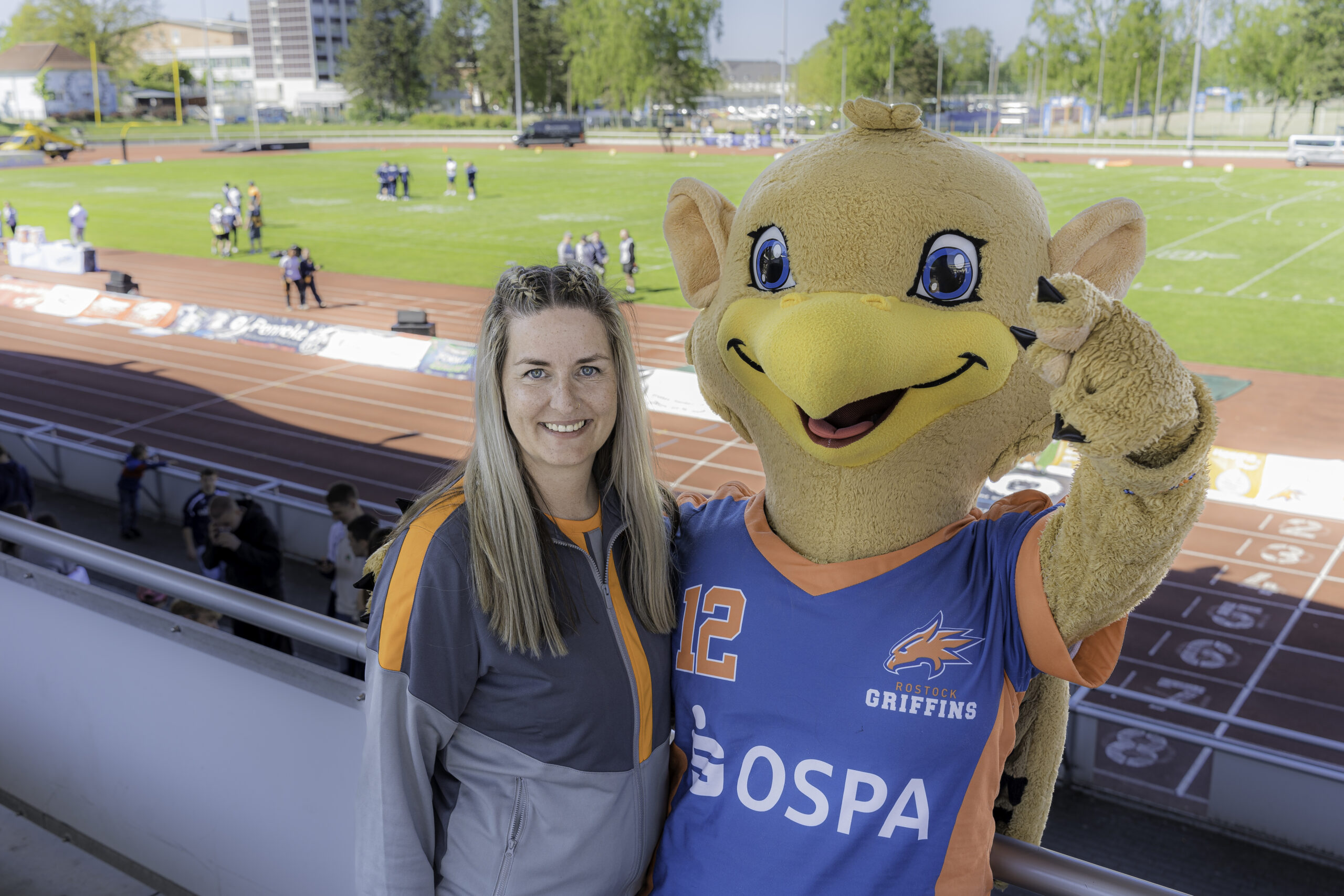 Peggy Schröder / Maskottchen Griffino. Peggy Schröder von den Rostocker Griffins, lange blonde Haare und blaue Augen, steht in Trainingszeug neben dem Vereinsmaskottchen Griffino - einem vogelartigen Fabelwesen mit rieseigen blauen Augen und einem gelben Schnabel.