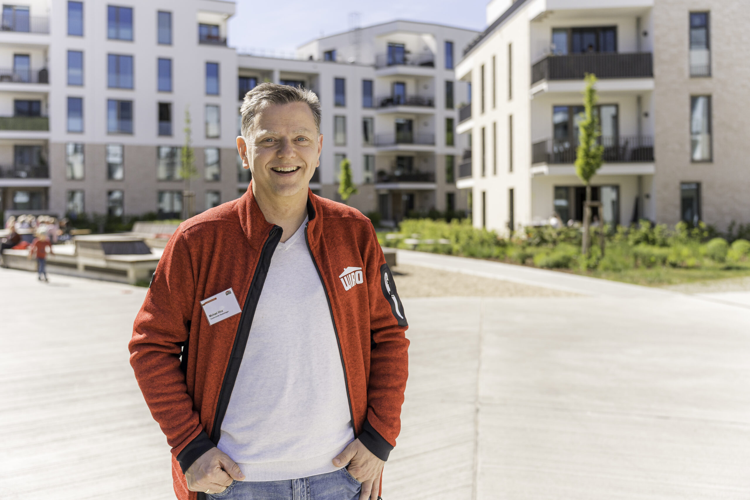 Michael Hinz (2) Michael Hinz trägt ein helles Shirt unter der roten WIRO-Jacke und strahlt in die Kamera. Im Hintergrund: Die Neubauten der Thierfelderstraße, für die er als Wohnungsverwalter verantwortlich ist.