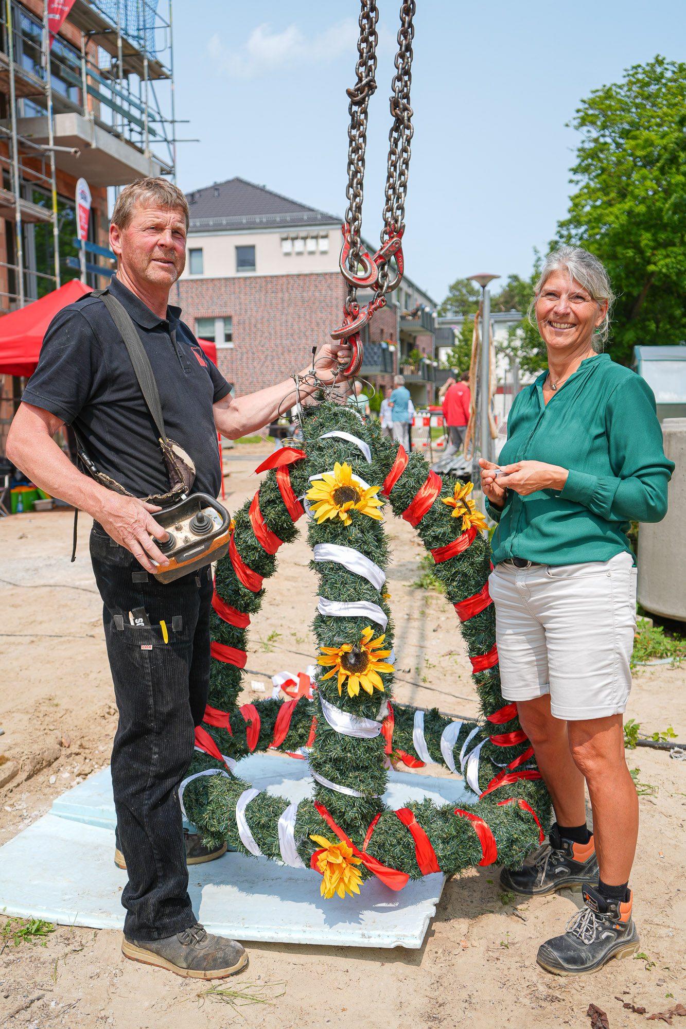 Richtfest Albin Köbis Straße Ein Handwerker in schwarzer Montur und Bauleiterin Kerstin Stunnack in grüner Bluse und weißen Shorts stehen neben der mit gelben Blumen und rot-weiß beschleiften grünen Richtkrone am Neubau in der Albin-Köbis-Straße in Markgrafenheide.