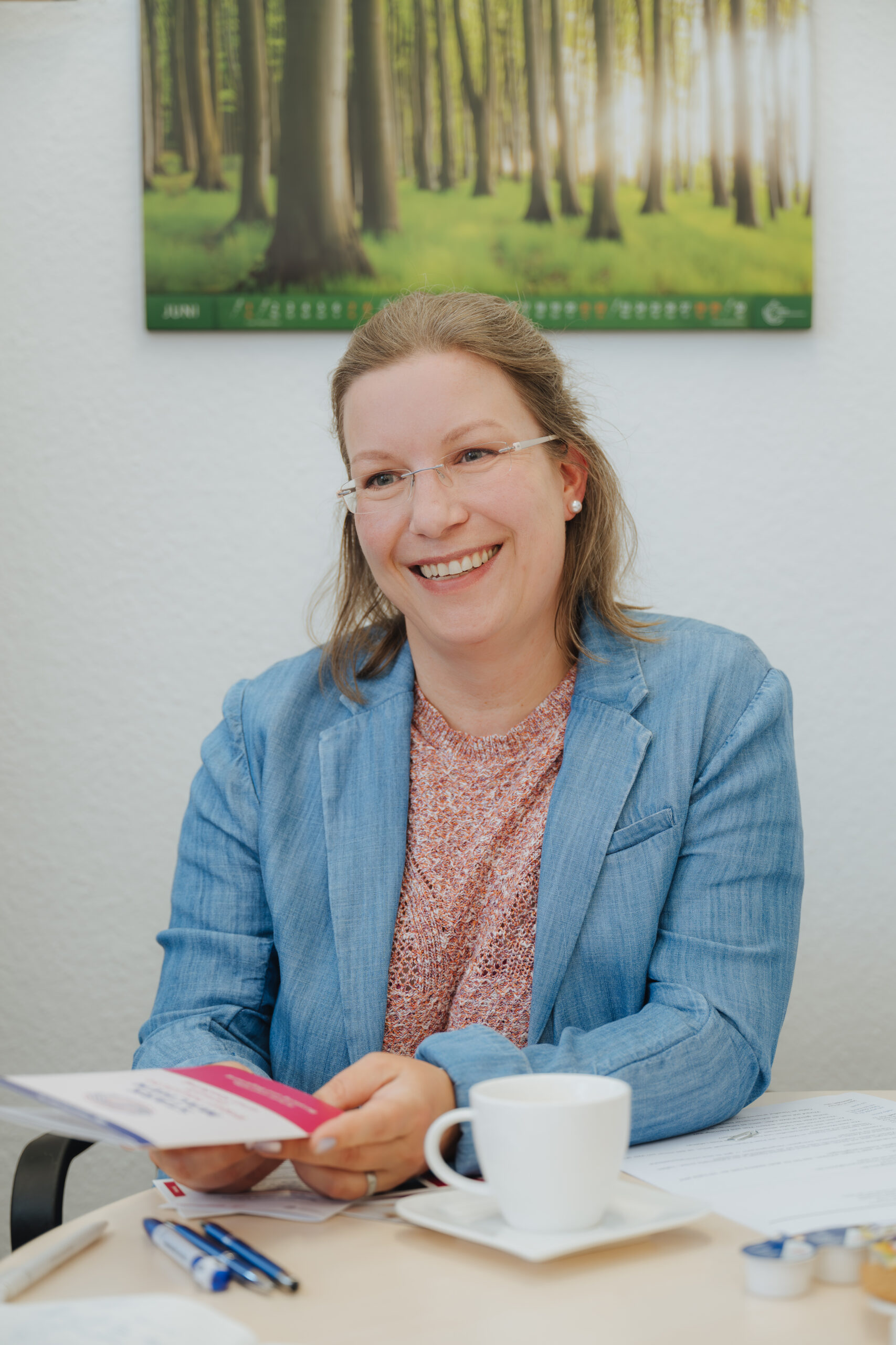 Kriminalhauptmeisterin Anke Symnazik Kriminalhauptmeisterin Anke Symnazik, hellbraune Haare, randlose Brille, sitzt im blauen Jeans-Blazer an einem Tisch, auf dem eine Kaffeetasse steht.