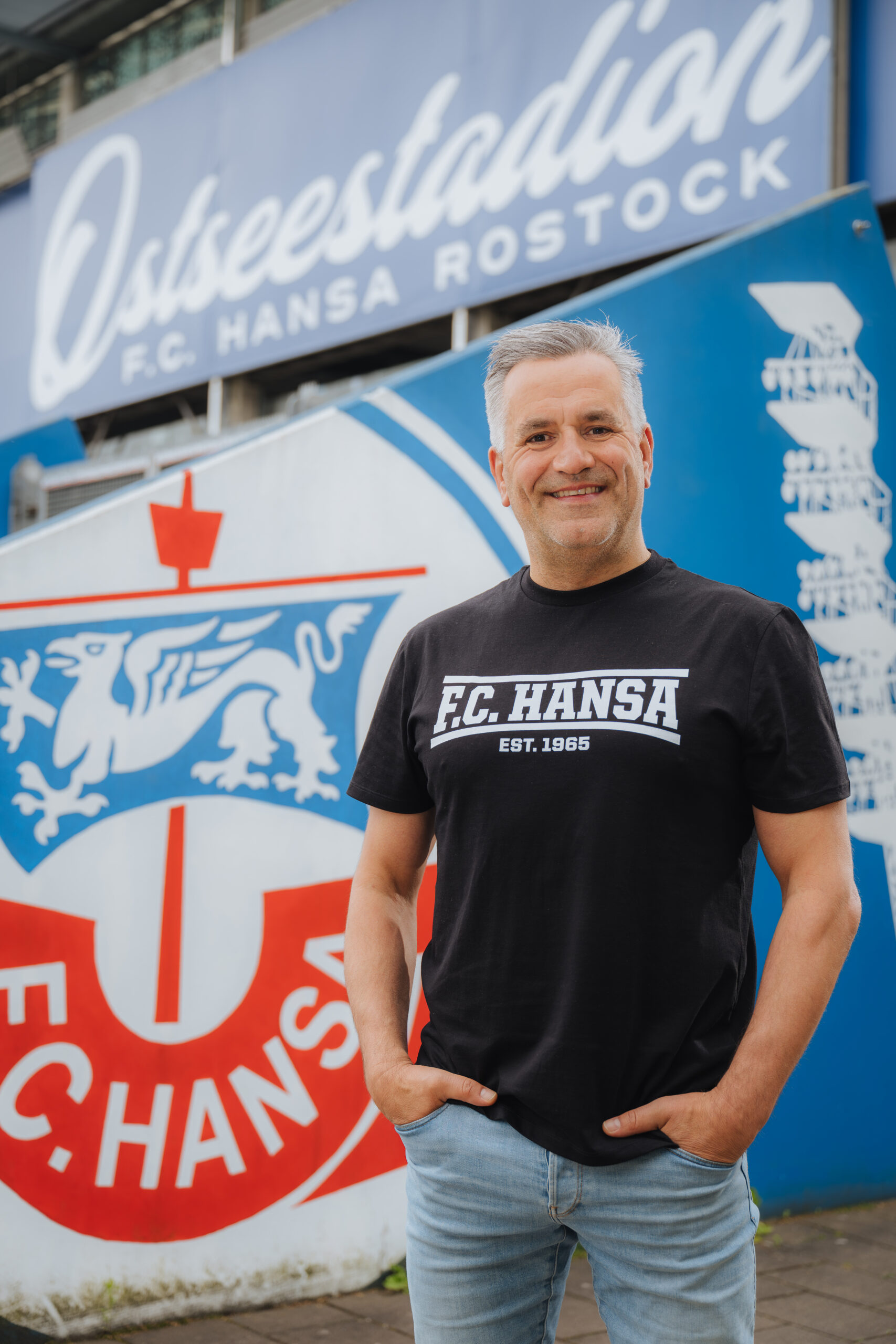 Michael Streu Michael Streu, graue kurze Haare, steht mit schwarzem F.C. Hansa-Shirt im Ostseestadion vor dem großen F.C. Hansa-Banner.