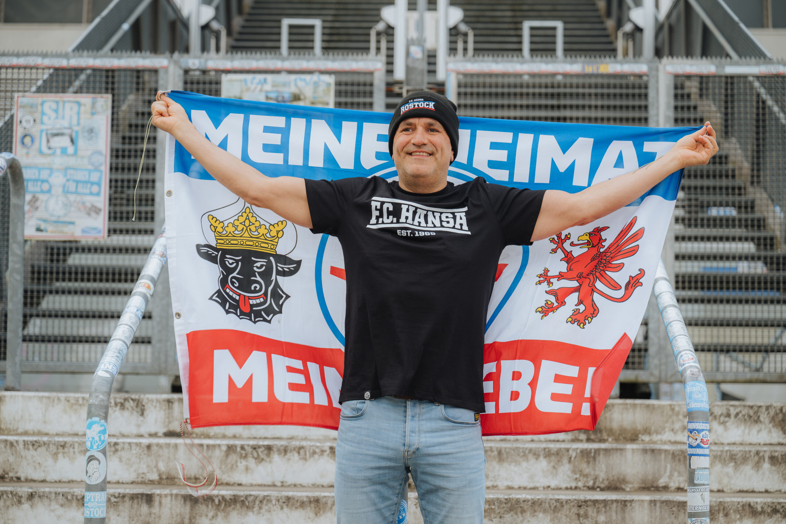 Michael Streu, F.C. Hansa-Fan der ersten Stunde steht im Ostseestadion mit schwarzer Mütze und schwarzem Shirt des F.C. Hansa und breitet die Club-Flagge hinter seinem Rücken aus. Auf der Flagge: Meine Heimat. Meine Liebe!