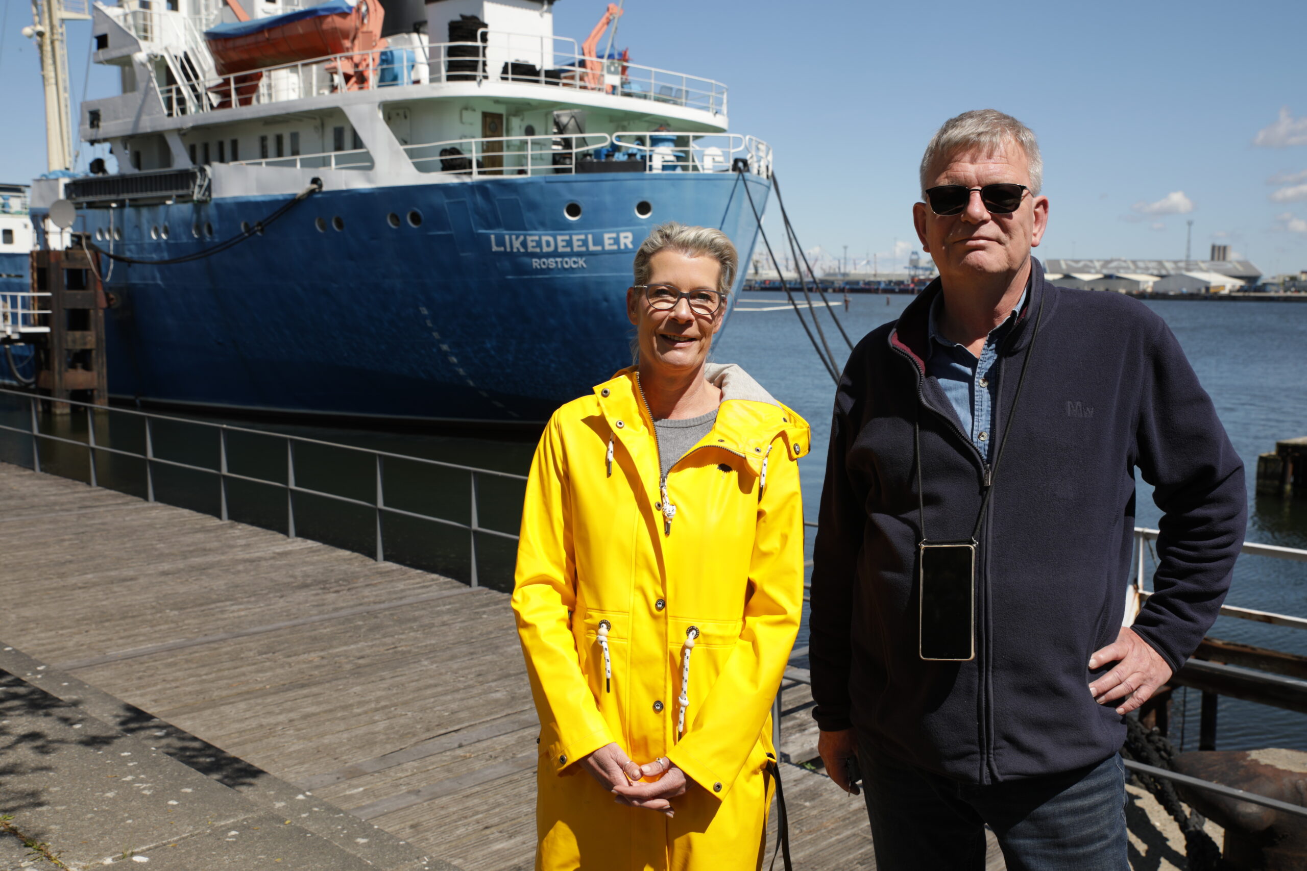 Taufe Vagel Grip Sigrid Bergholz im gelben Regenmantel, zuständig für die Kinder- und Jugendarbeit, und Peter Neumann, der eine dunkle Jacke und eine Sonnenbrille trägt und der Geschäftsführer und Projektkoordinator des Fördervereins Likedeeler ist, stehen vor dem Jugendschiff Likedeeler.