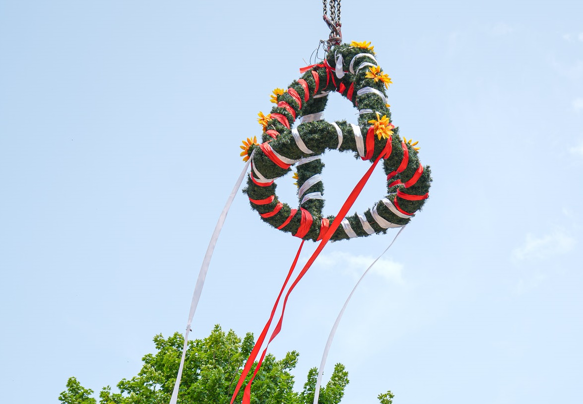 Die begrünte und mit gelben Blumen und rot-weißen Schleifen geschmückte Richtkrone der Albin Köbis-Straße in Markgrafenheide schwebt in luftiger Höhe.