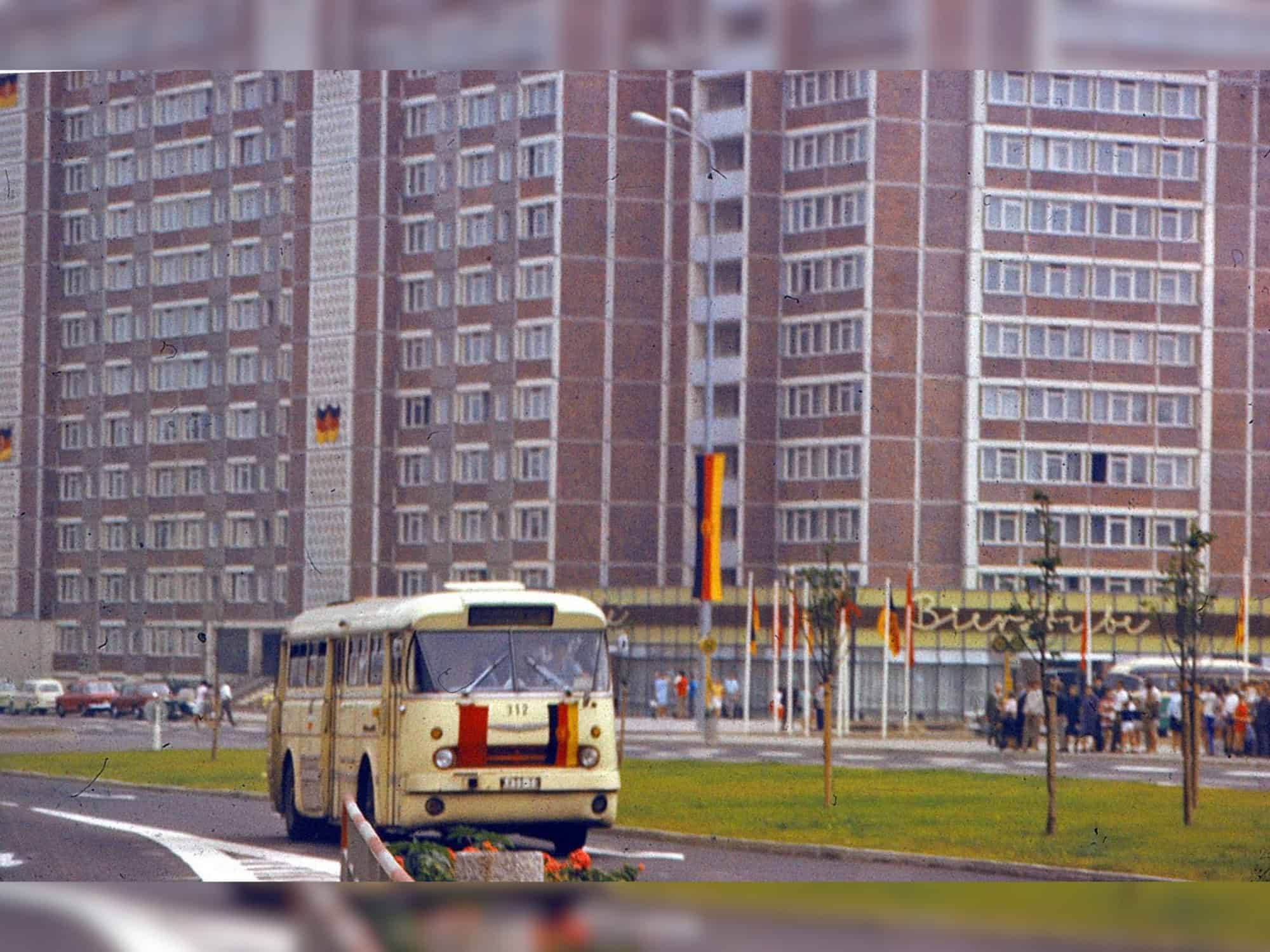 Historische Aufnahmen Hochhaus Am Vögenteich Vor dem Hochhaus Am Vögenteich fährt ein gelber DDR-Bus, geschmückt wie das Haus selbst, mit DDR-Fahnen. Am Haus gibt es ein Cafe und eine Bierstube.