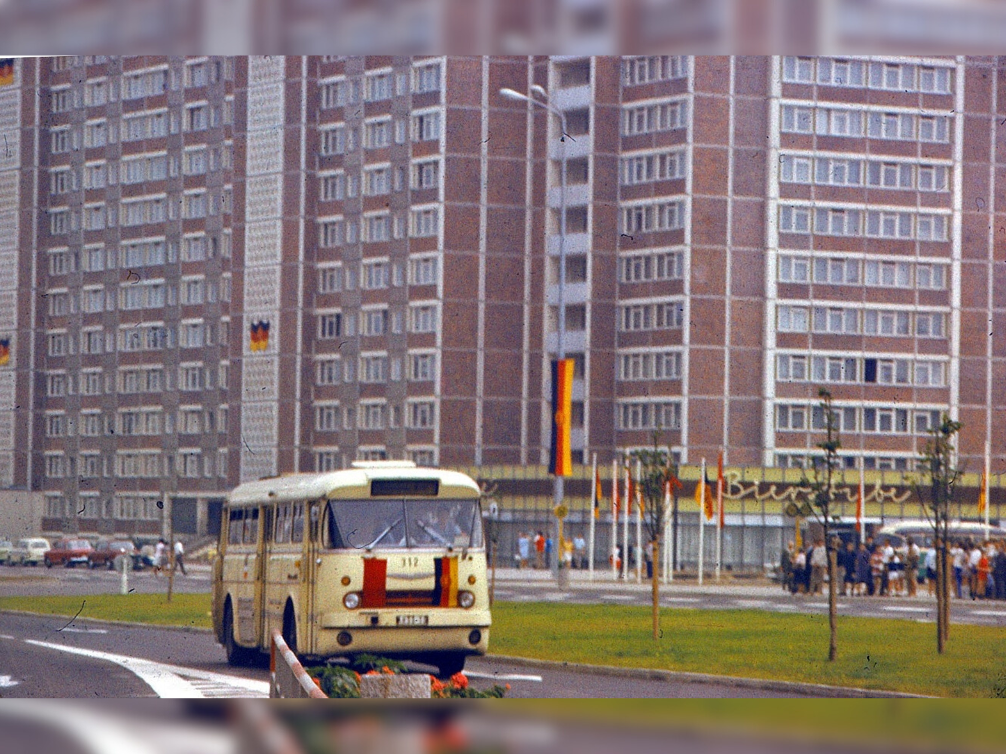 Historische Aufnahmen Hochhaus Am Vögenteich Vor dem Hochhaus Am Vögenteich fährt ein gelber DDR-Bus, geschmückt wie das Haus selbst, mit DDR-Fahnen. Am Haus gibt es ein Cafe und eine Bierstube.