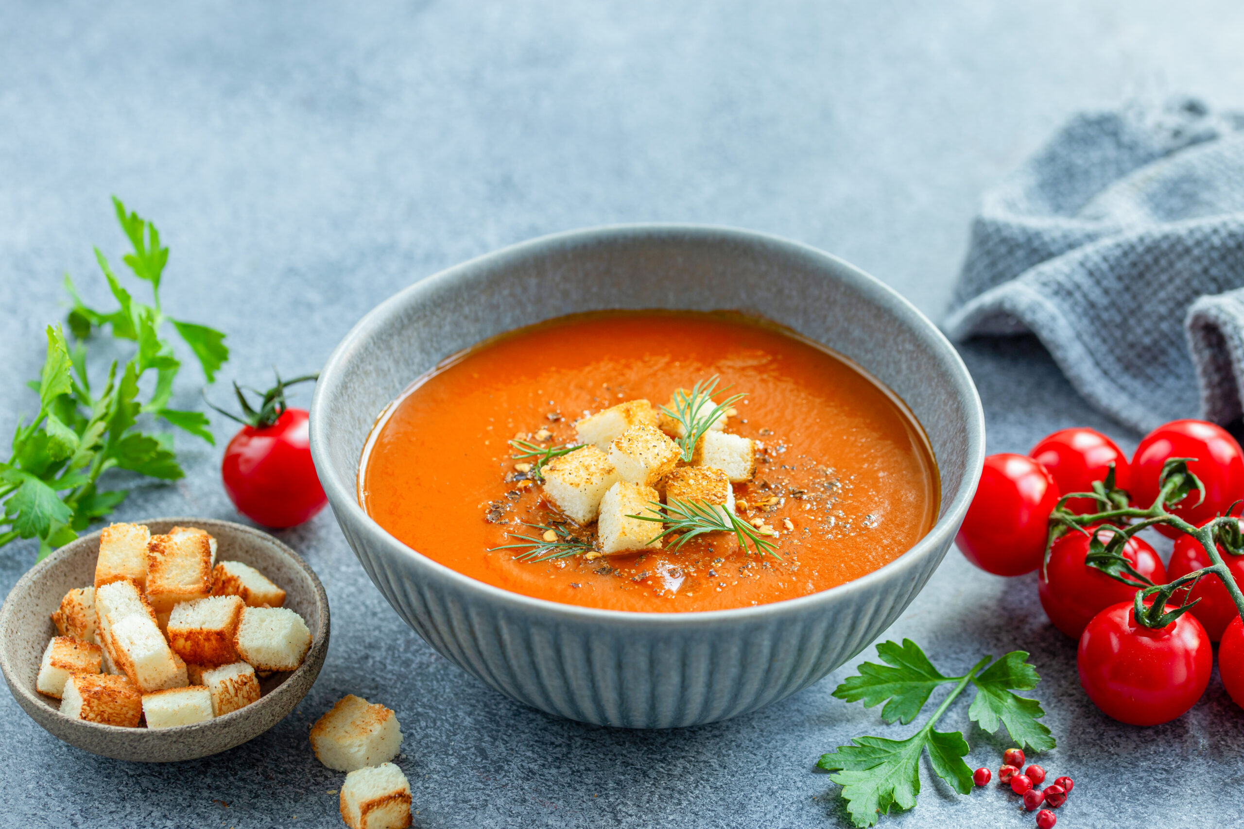 Eine graue Schale mit Gazpacho, daneben Tomaten, grüne Kräuter und knusprig braue Croutons.