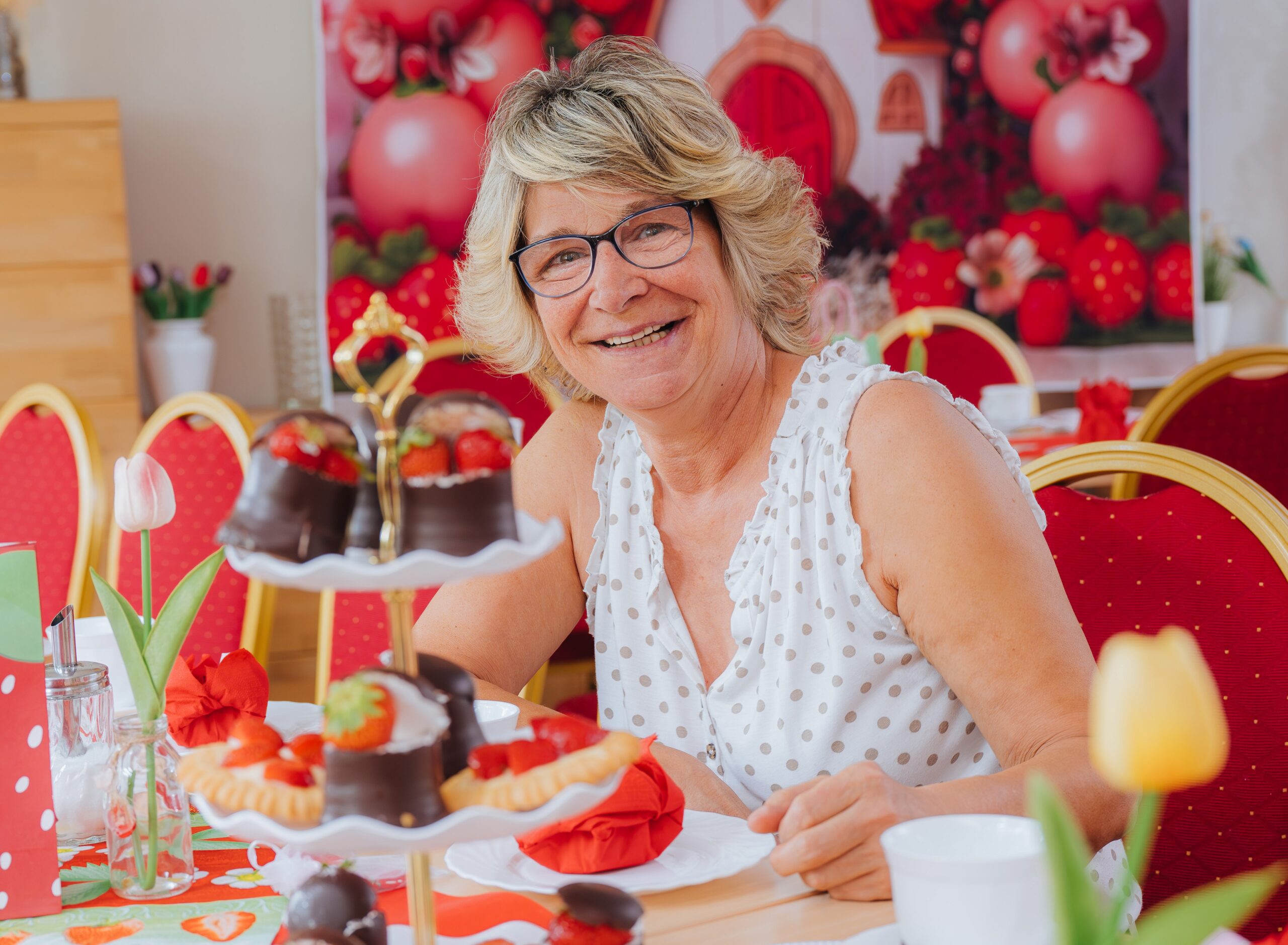 Katrin Braecker von der Begegnungsstätte der Volkssolidarität ist immer für Erdbeeren und Spaß zu haben. Um sie herum Erdbeeren auf Etageren, Erdbeeren auf einem Bild und Stühle in Erdbeerfarbe.