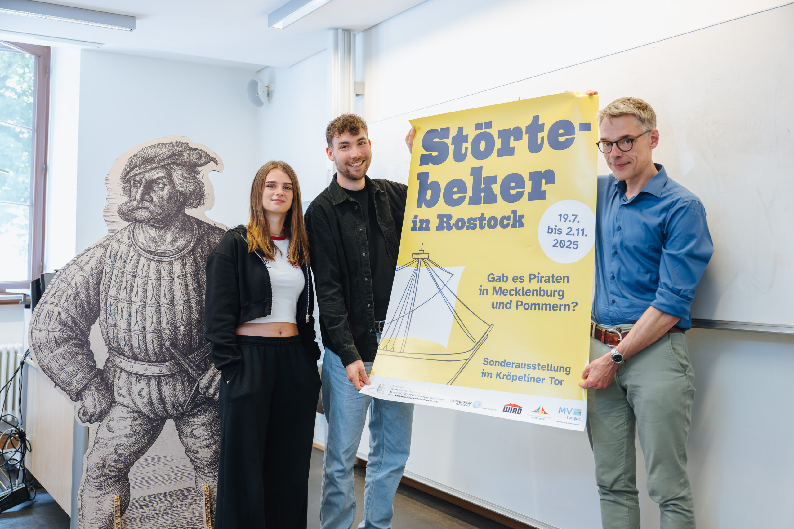 Professor Gregor Rohmann (re.), Julian Max Wahle und Ellena Ditscher, beide Studenten, neben einem lebensgroßen Aufsteller von Klaus Störtebeker.