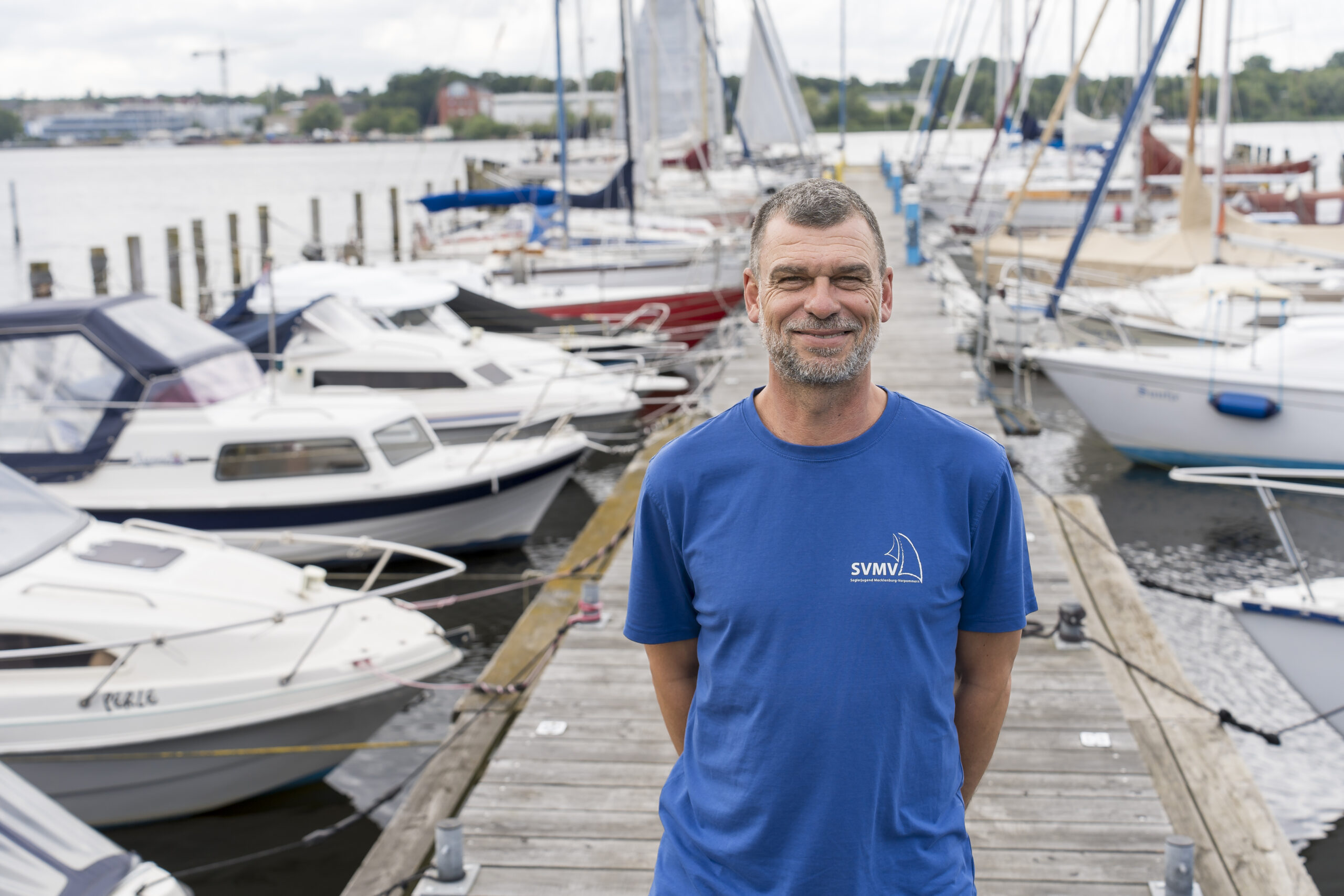 Frank Hildebrandt, graue Haare, grauer Bart mit breitem Grinsen im Gesicht am Steg vom Yachtclub Warnow, an dem viele Segelboote anliegen.