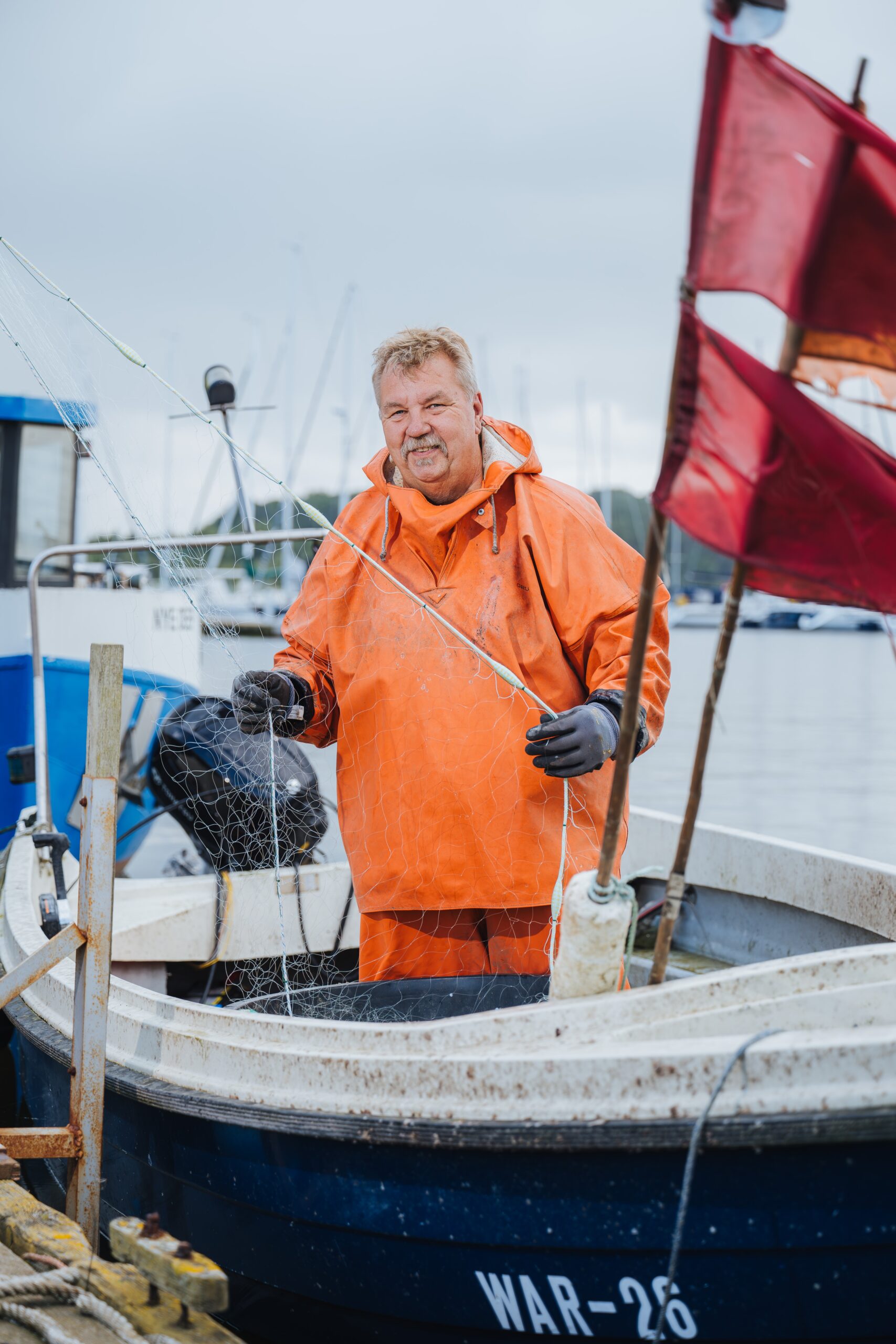 Fischer Torsten Rudolph im orange-farbigen Ölzeug und einer Angelrute in der Hand auf seinem Kutter am Schmarler Ufer.