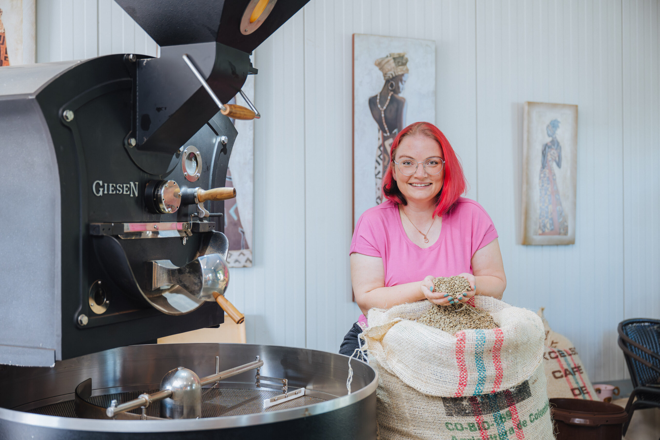 Kristine Rönnpagel schöpft mit Ihren Hände rohe Kaffeebohnen aus einem großen Kaffeesack. Sie steht neben einer großen Röstmaschine.