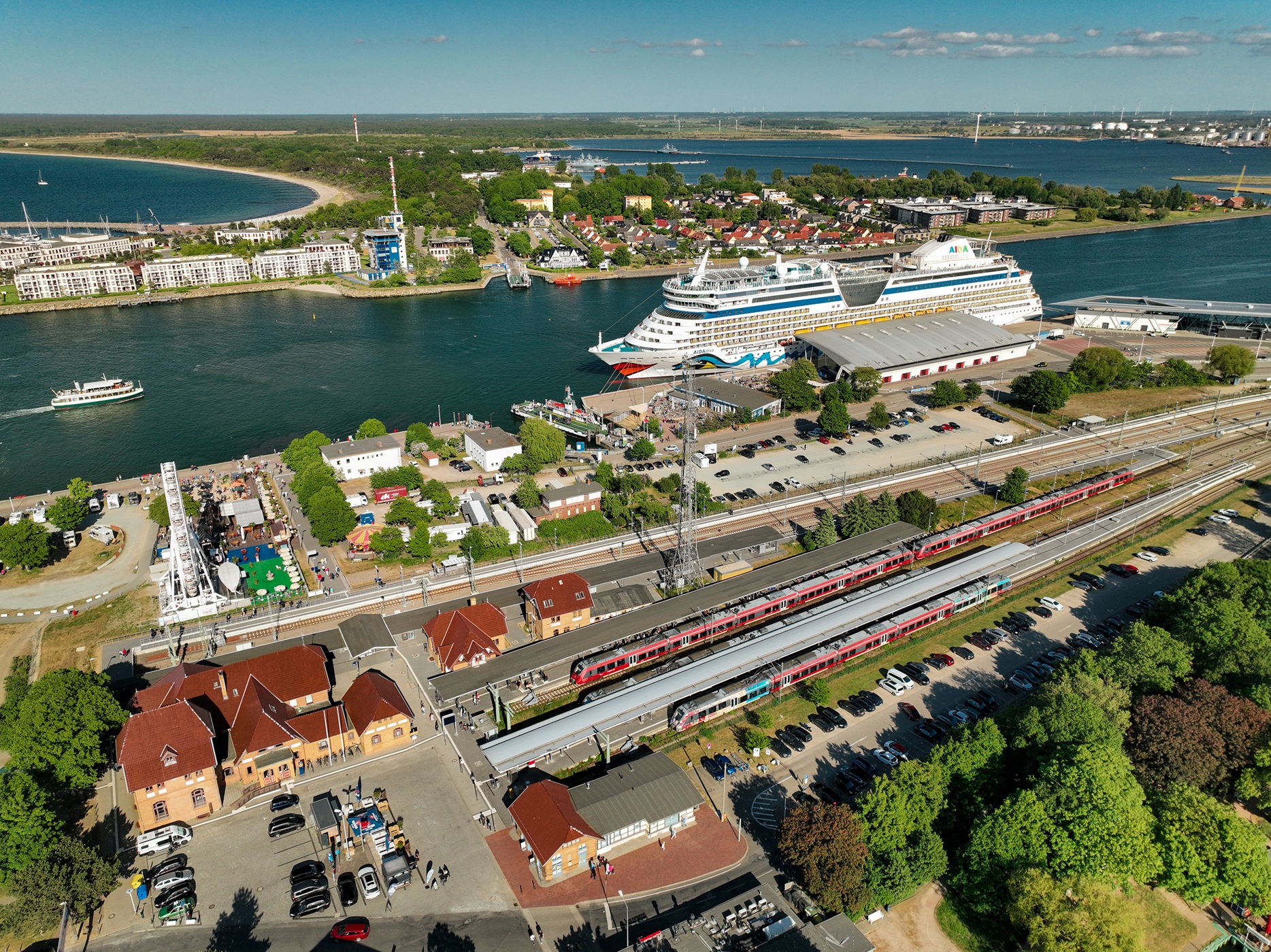 Luftaufnahme mit dem Parkplatz Am Bahnhof in Warnemünde. Einfahrende Züge steuern Richtung Bahnhof. Im Hintergrund liegt ein Kreuzfahrtschiff am Fähranleger Richtung Ostsee.