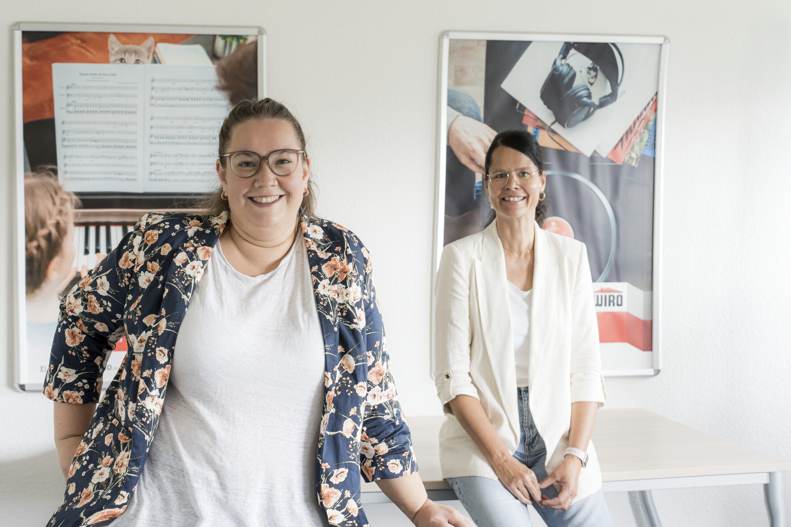 Nina Schoenfeld (links), Pferdeschwanz, Brille und großes Lachen über dem geblümten Blazer, und Manuela Nimmich, dunkler Zopf, dezente Brille und weißer Blazer über Jeans, sitzen vor WIRO-Plakaten und wissen alles zum Thema Nachmieter für WIRO-Wohnungen.