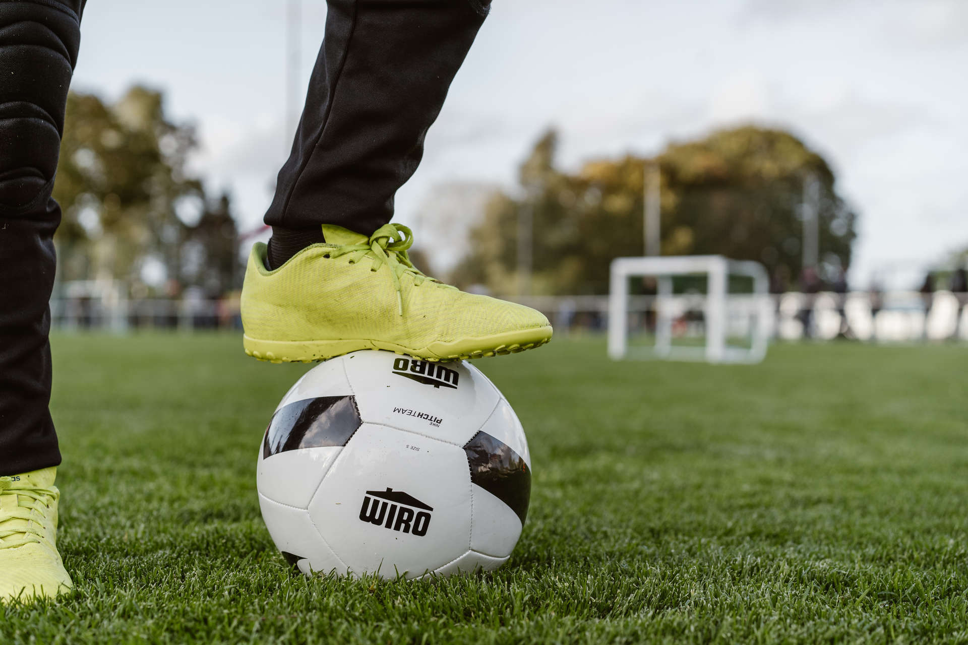 Ein Kunstrasenplatz. auf dem ein schwarz-weißer Fußball mit WIRO-Logo liegt, auf dem sich ein Bein in schwarzen Sporthosen und gelben Turnschuhen stützt.