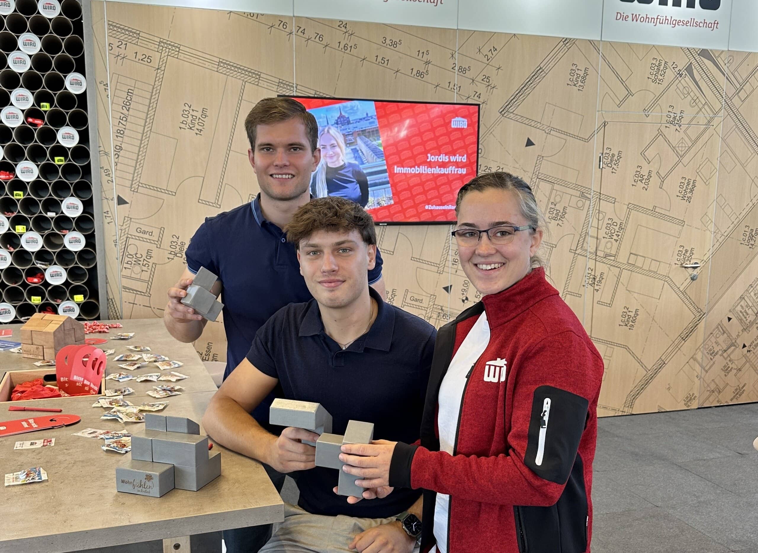 Drei junge Azubis, zwei davon Männer in dungelblauen Poloshirt, am Messestand der WIRO auf der Jobfactory. Die einzige Dame im Bunde trägt eine rote WIRO-Jacke mit weißem WIRO-Logo.