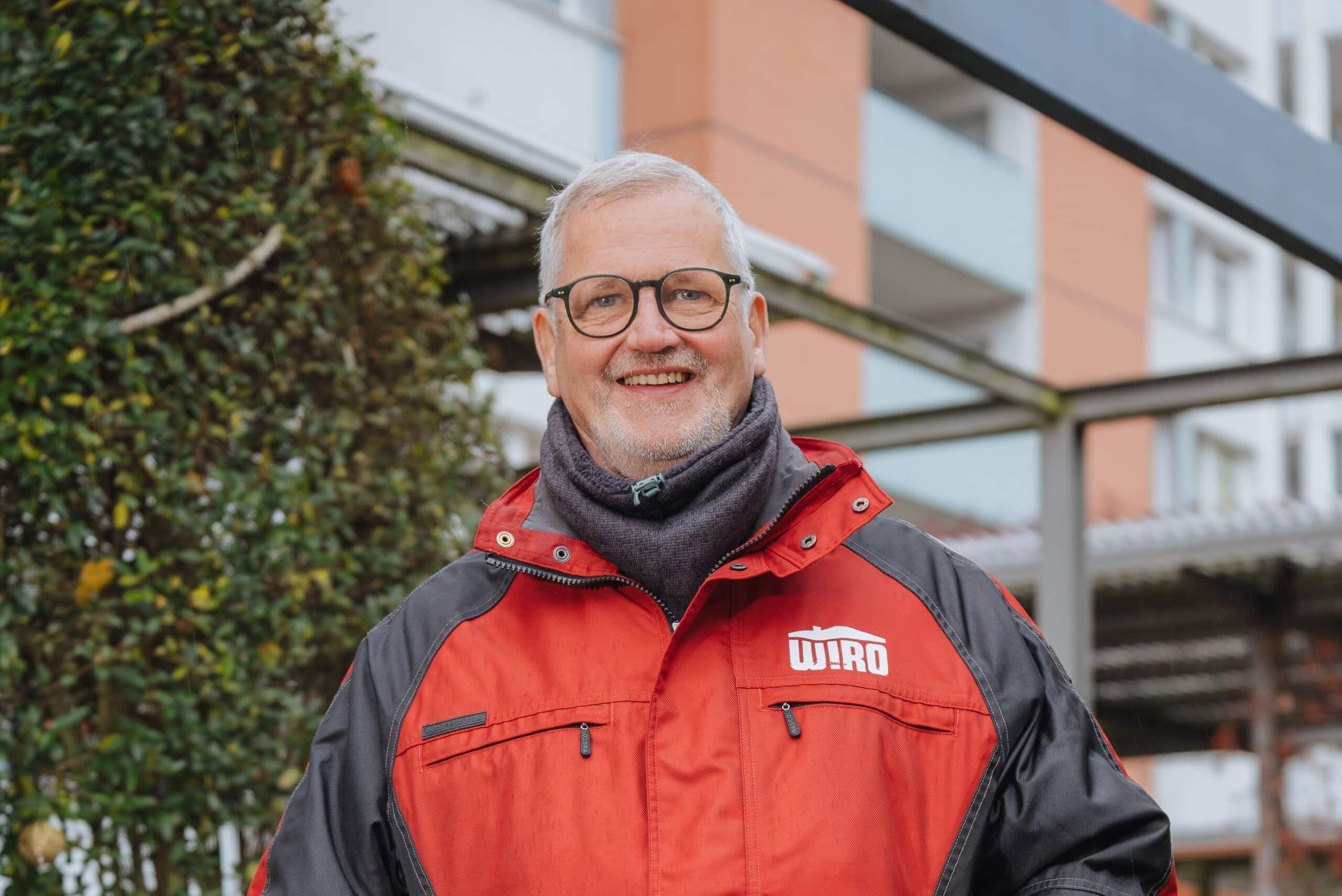 Portrait vom WIRO-Hausmeister Michael Röllig mit weißen kurzen Haaren, dunkler Brille und in roter Jacke.