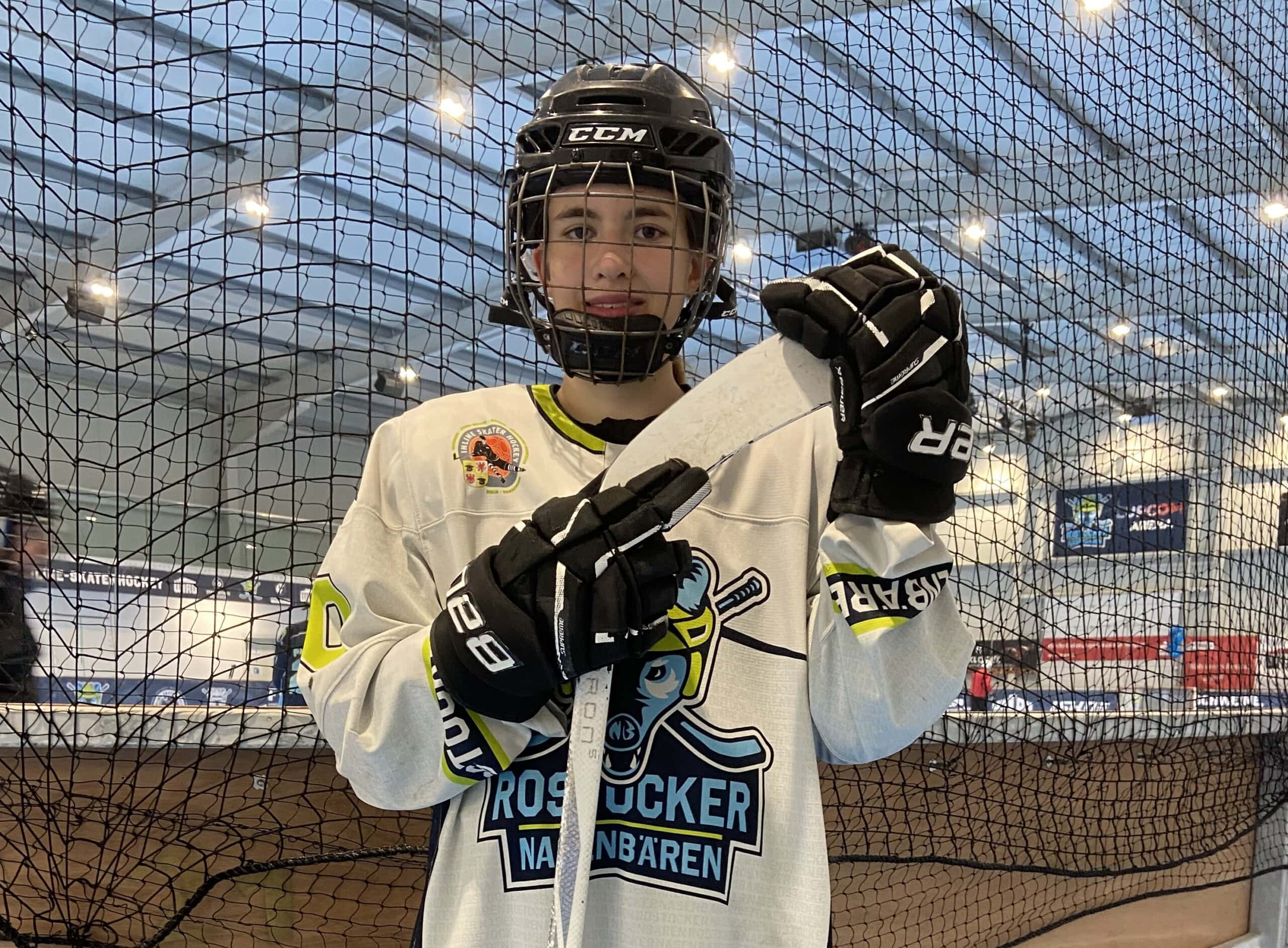 Ein junger Spieler der Inlinehockey-Mannschaft Rostocker Nasenbären mit Helm, weißem Shirt und blauem Logo. In den dick behandschuhten Händen hält er einen Schläger.