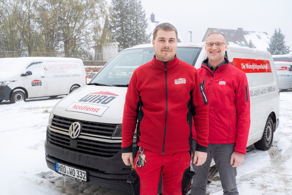 Zwei Kollegen vom WIRO-Bauservice in roter Arbeitsmontur vor einem WIRO-Transporter mit der Aufschrift Notdienst auf dem verschneiten Bauhof.