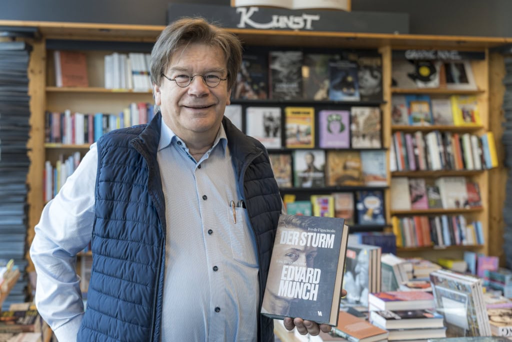 Manfred Keiper, Inhaber der anderen Buchhandlung am Doberaner Platz, mit Lesebrille und einem Buch in der Hand vor einem Bücherregal.