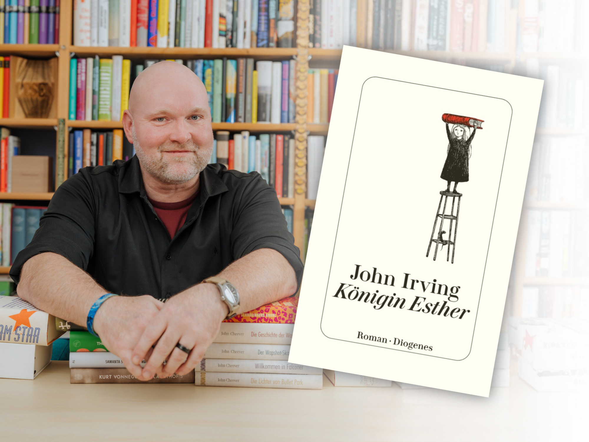 Bookster Stefan Härtel sitzt vor einem großen Bücherregal stützt seine Arme auf Bücherstapel, neben ihm das Titelbild des Romans "Königin Esther" von John Irving