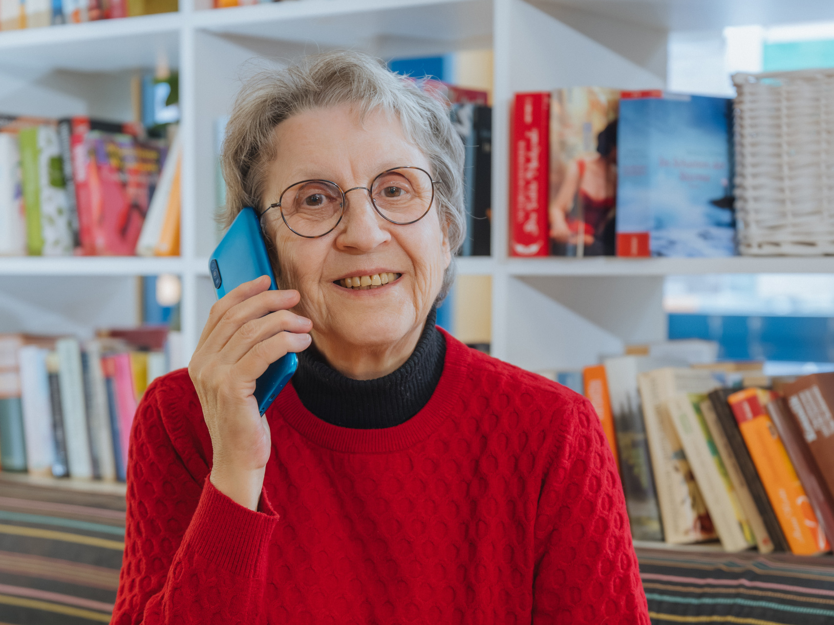 Patricia Fleischer sitzt mit einem roten Pullover auf einem Sofa und hält ein Telefon in der Hand.