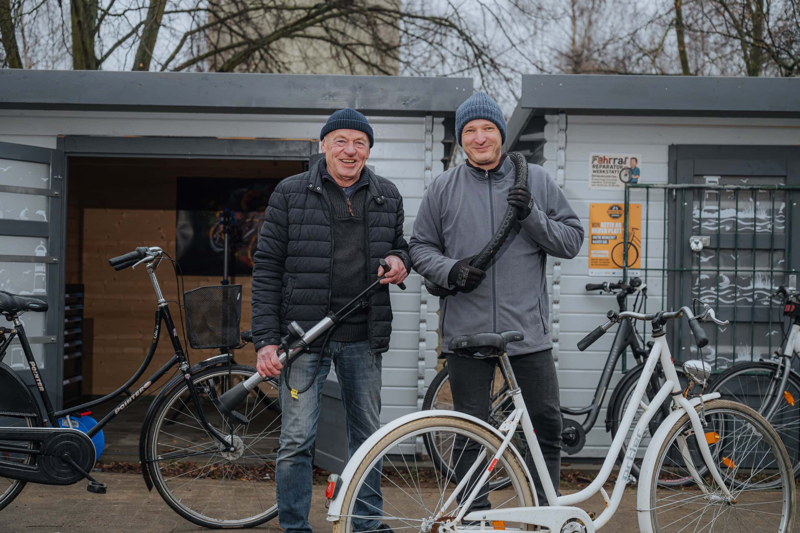 Ehrenamtlich schrauben Siegfried Kirchner und Henryk Henschke mit Schülern in der Fahrradwerkstatt der Rostocker Nordlichtschule. Im Bild stehen die beiden Männer zwischen mehreren Fahrrädern und halten Schläuche und Luftpumpen in den Händen.