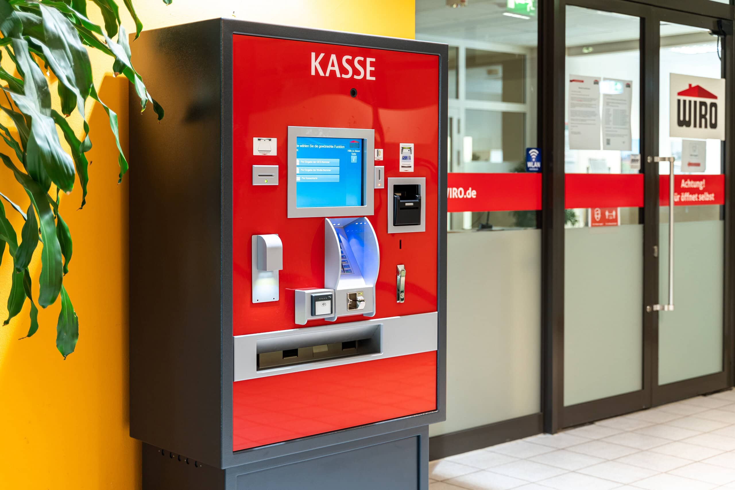 Ein roter Kassenautomat der WIRO neben der im WIRO-Design gestalteten verglasten Eingangstür eines KundenCenters.