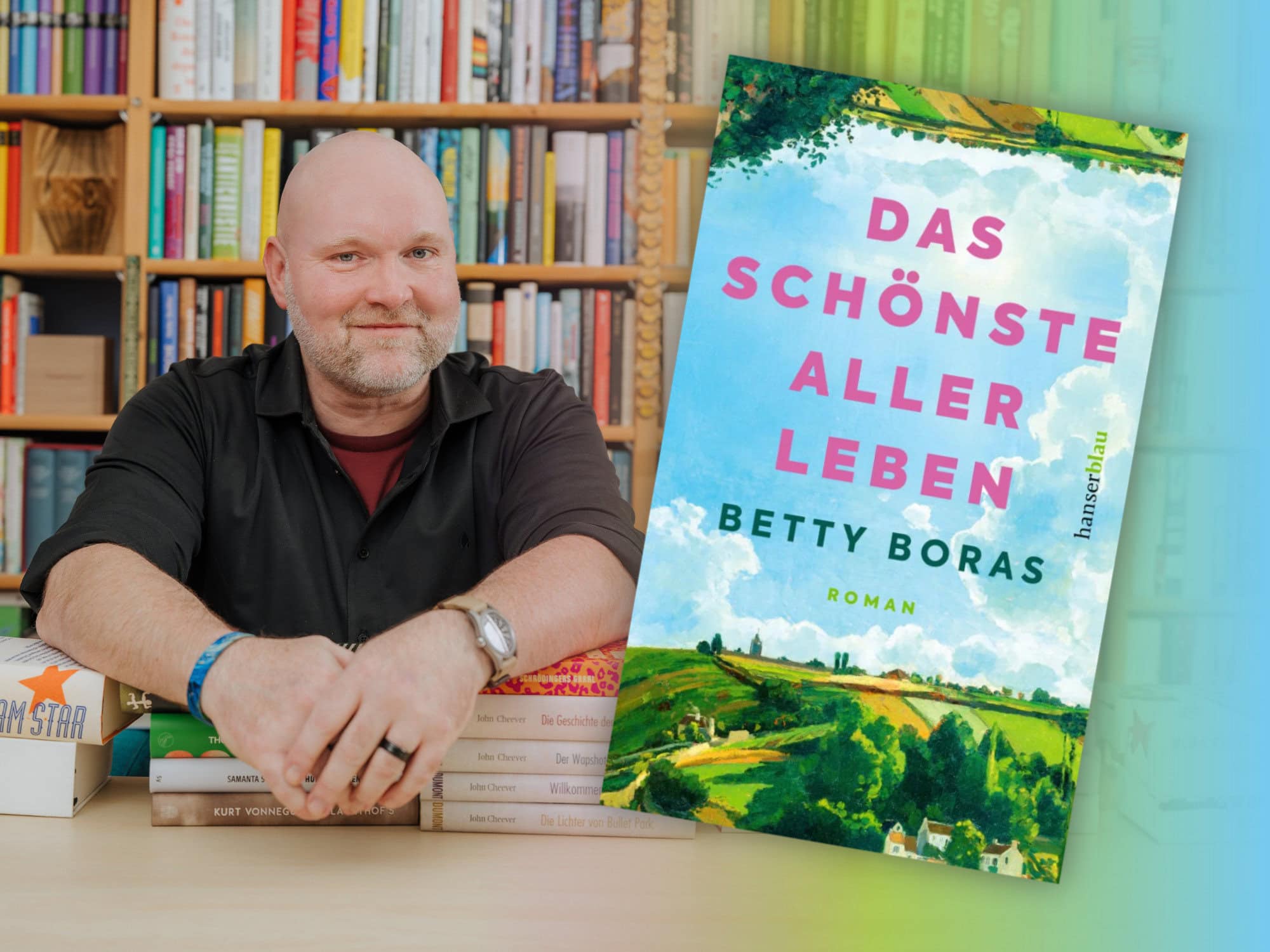Bookster Andreas Härtel sitzt vor einem großen Bücherregal stützt seine Arme auf Bücherstapel, neben ihm das Titelbild des Romans "Das schönste aller Leben" von Betty Boras