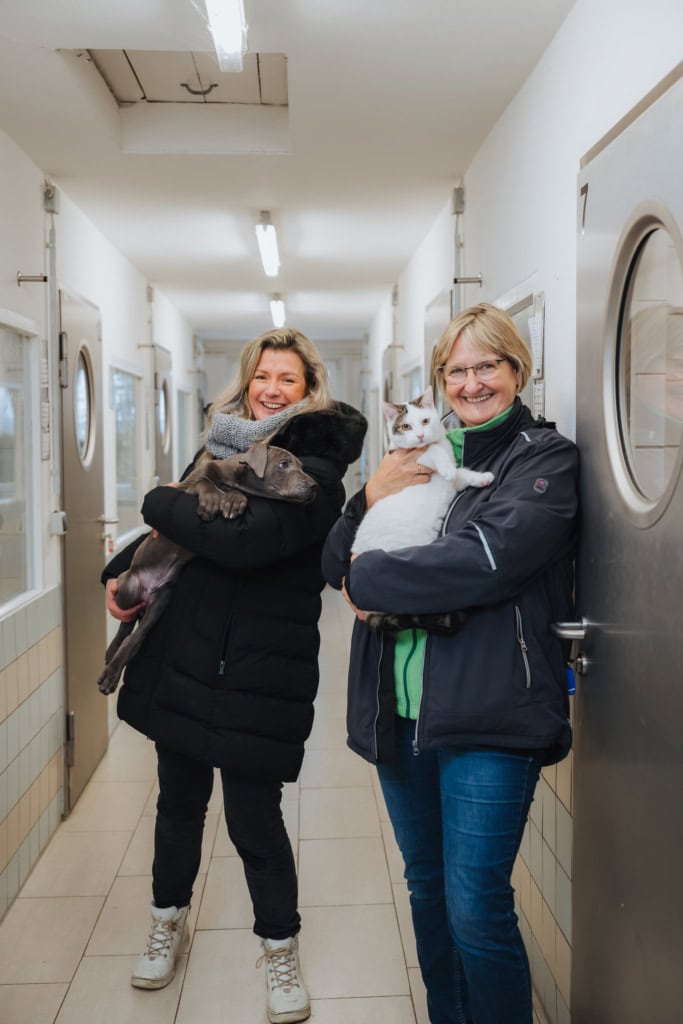 Angelika Streubel (li.), Tierheim-Mitarbeiterin und Vorsitzende des Rostocker Tierschutzvereins, mit Leiterin Regina Groß. Angelika hält einen Welpen, Regina eine Katze auf dem Arm. Der Welpe zeigt deutlich Interesse an der Katzen-Mitbewohnerin im Tierheim Schlage.
