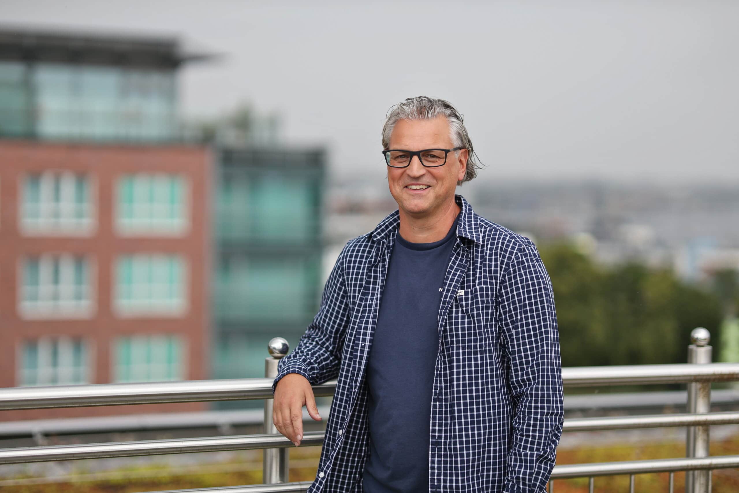 Silvio Krüger ist bei der WIRO zuständig für die Betriebskostenabrechnungen. Er steht mit zurück gekämmten grauen Haaren, dunkler Brille und weiß-dunkel kariertem Hemd auf der Terrasse des WIRO-Hauptgebäudes.