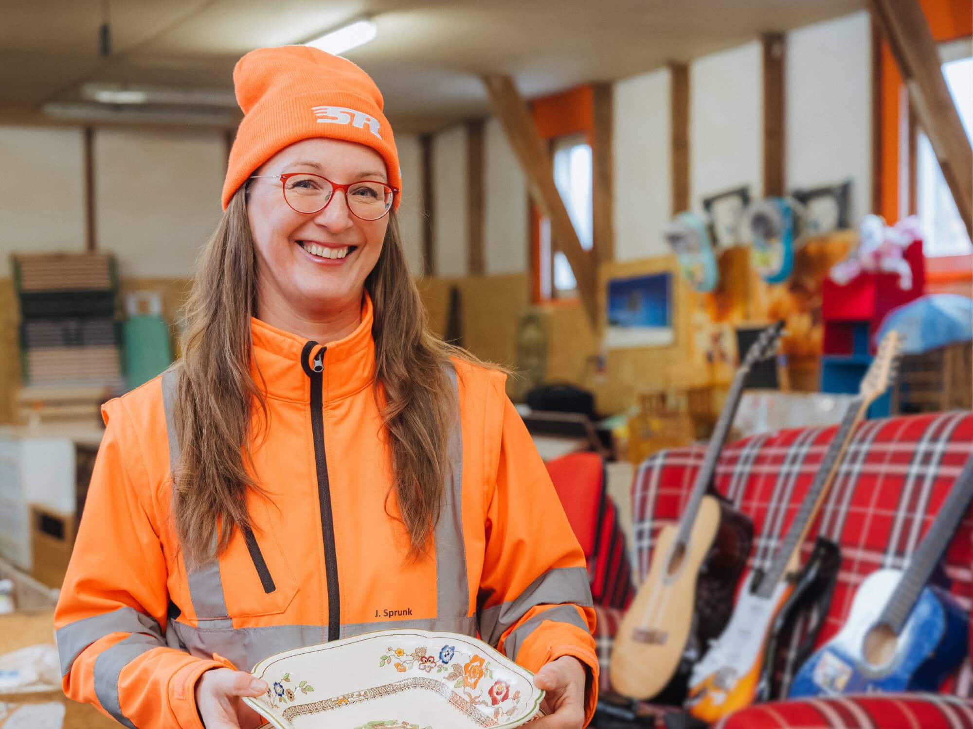 Leiterin des Rückkonsumzentrums Hanseschatz Juliane Sprunk, langes Haar, orange Kleidung und Mütze, hält eine Porzellan-Schale und steht vor den Artikeln des Hanseschatz-Angebotes