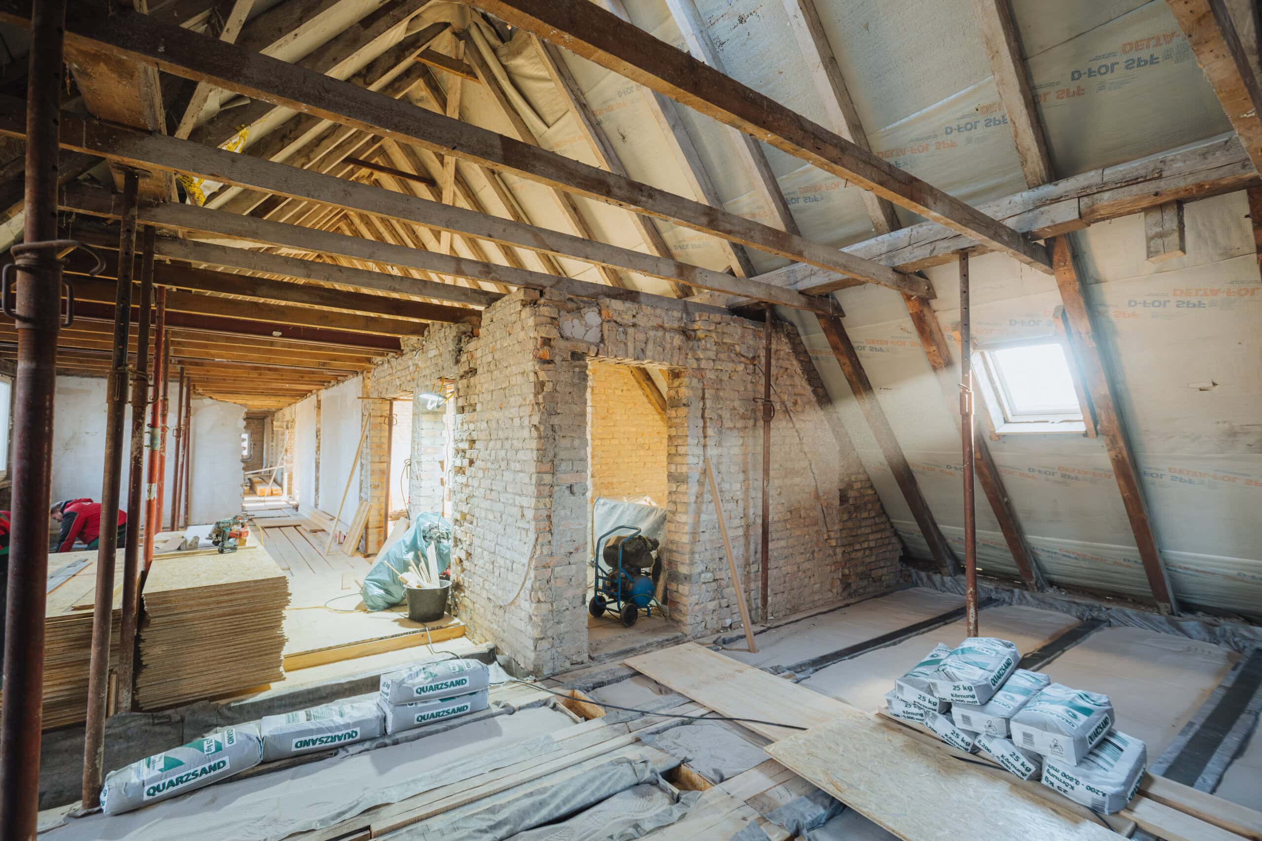 Zementsäcke liegen auf dem Boden, Holzplatten sind gestapelt. Die Wände haben keinen Putz mehr, die Balken sind frei gelegt.