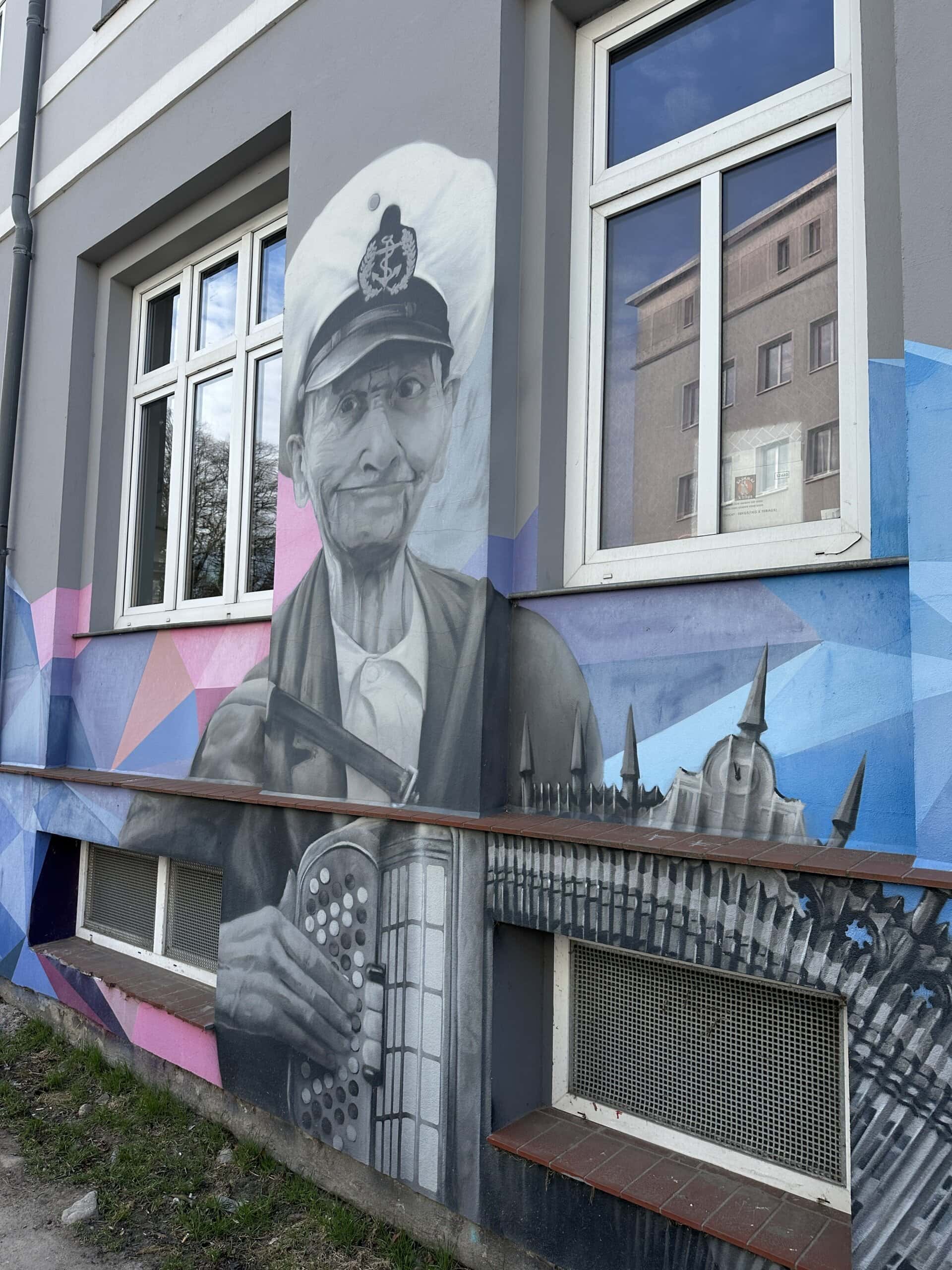 Ein alter Mann mit Kapitänsmütze und einem Akkordeon in der Hand ist als Graffiti auf eine Hauswand gesprüht.