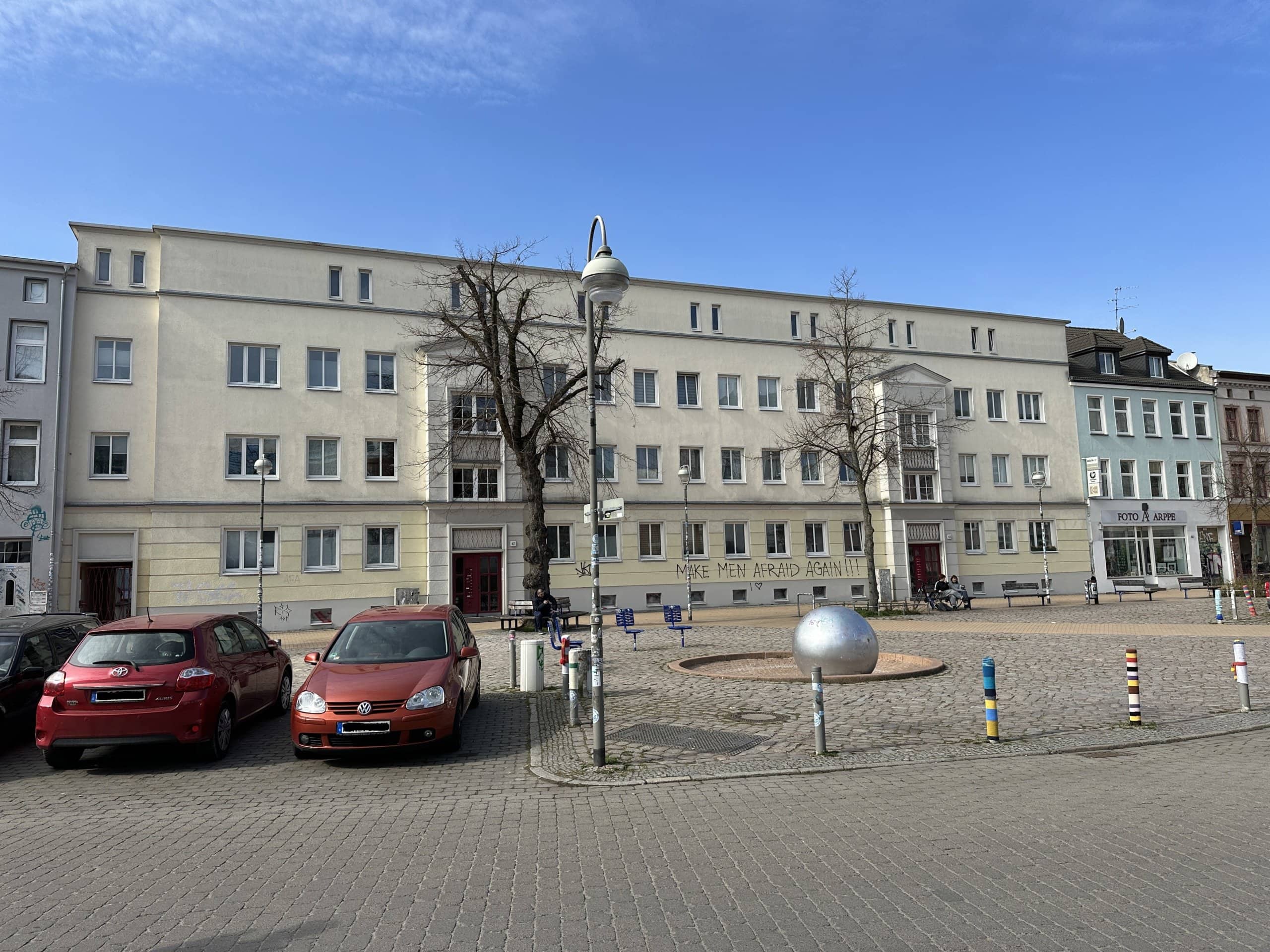Ein großes Mehrfamilienhaus in Rostock. Die Fassade ist gelblich. Davor sitzen Menschen auf einer Bank.