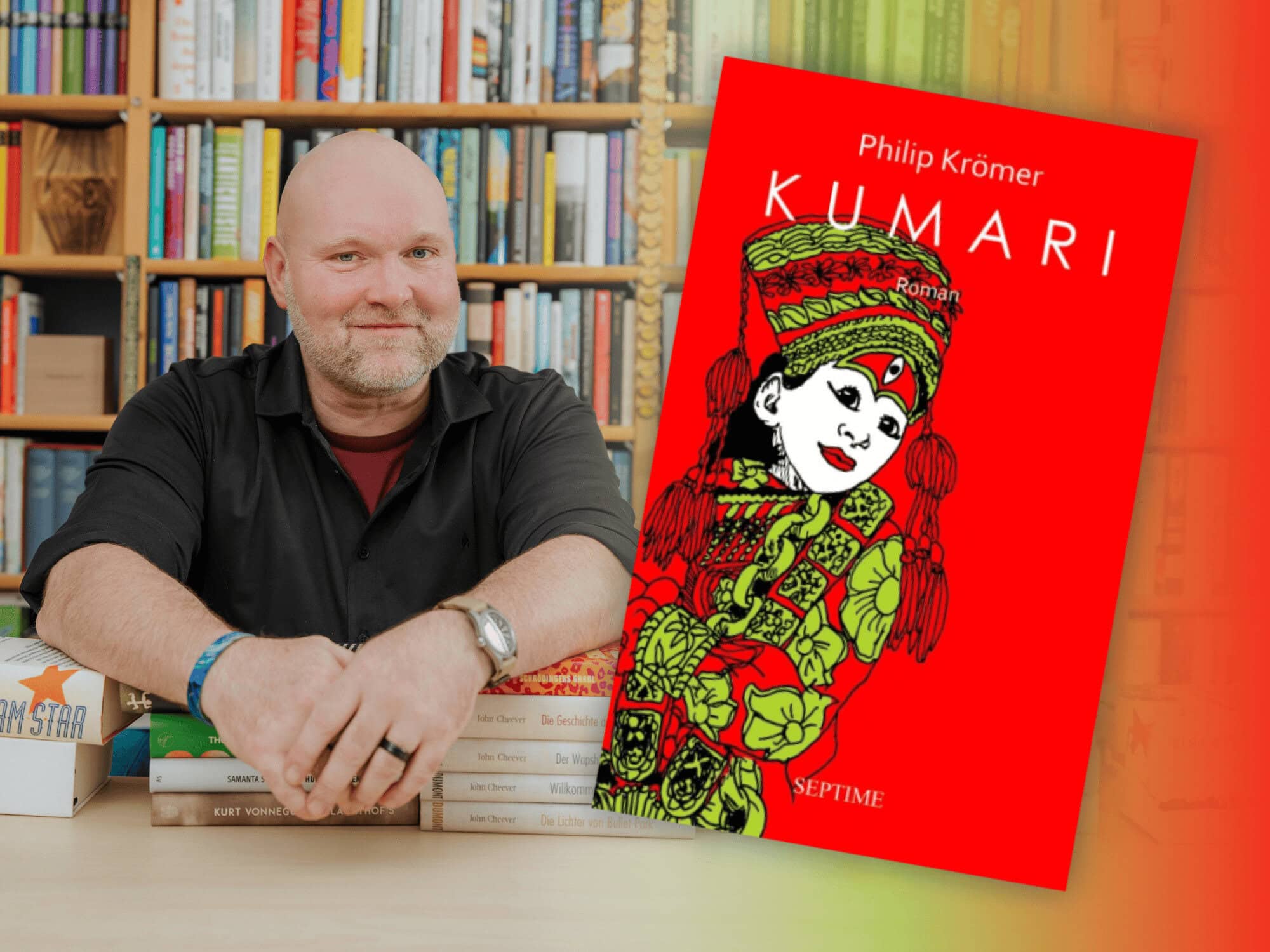 Bookster Stefan Härtel sitzt vor einem großen Bücherregal stützt seine Arme auf Bücherstapel, neben ihm das Titelbild des Romans "Kumari" von Philip Krömer
