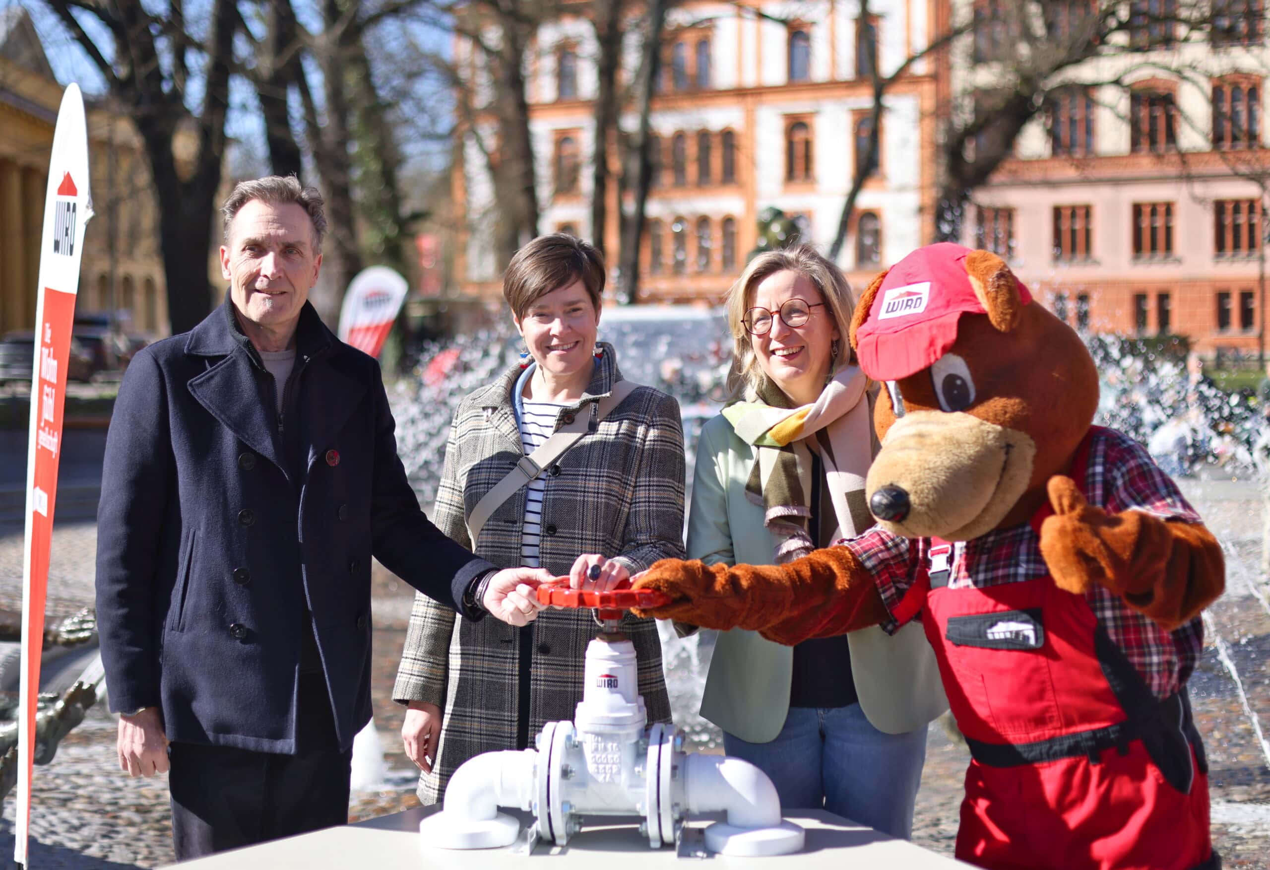 Bei der Rostocker Brunneneröffnung 2026 - Im Hintergrund der "Brunnen der Lebensfreude", davor von links nach rechts: WIRO-Geschäftsführer Ralf Zimlich, Oberbürgermeisterin Eva-Maria Kröger, Senatorin Dr. Ute Fischer-Gäde, WIRO-Maskottchen Bodo drehen gemeinsam den Brunnen an (Foto: WIRO/Danny Gohlke)
