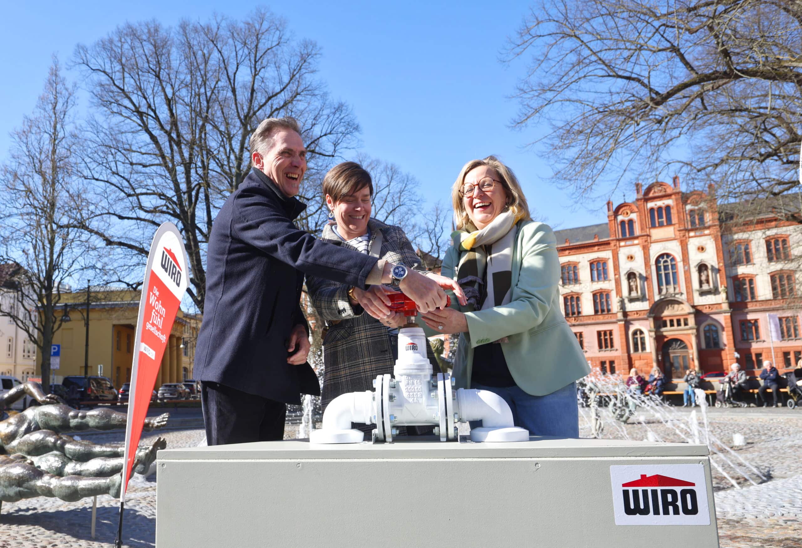 Bei der Rostocker Brunneneröffnung 2026 - Im Hintergrund der "Brunnen der Lebensfreude", davor von links nach rechts: WIRO-Geschäftsführer Ralf Zimlich, Oberbürgermeisterin Eva-Maria Kröger, Senatorin Dr. Ute Fischer-Gäde, WIRO-Maskottchen Bodo drehen gemeinsam den Brunnen an (Foto: WIRO/Danny Gohlke)
