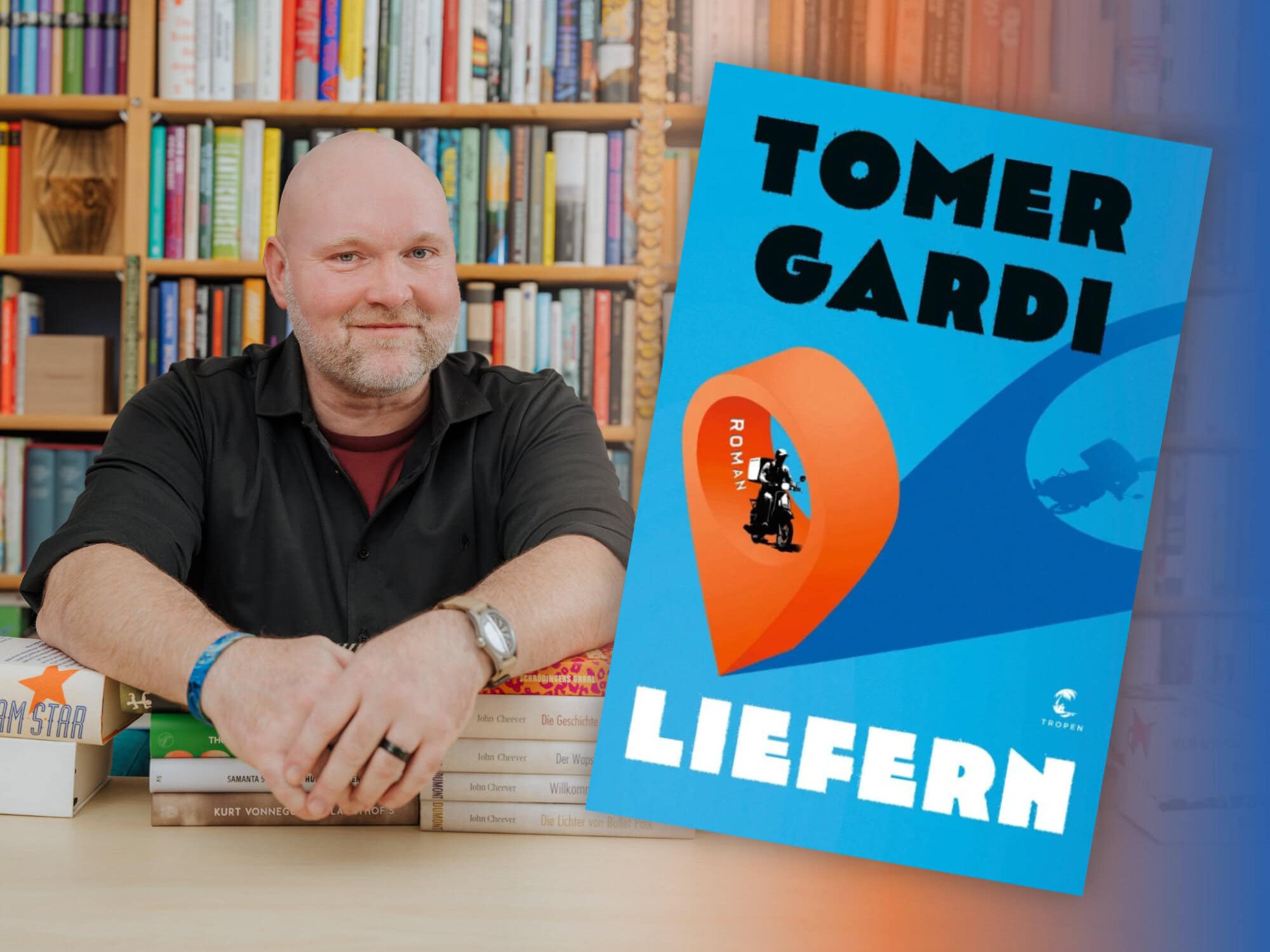 Bookster Stefan Härtel sitzt vor einem großen Bücherregal stützt seine Arme auf Bücherstapel, neben ihm das Titelbild des Romans "Liefern" von Tomer Gardi